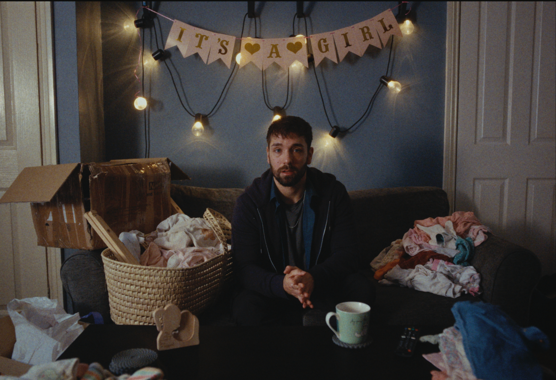 A man with dark hair and a beard, wearing a dark jacket and sitting on a cluttered couch surrounded by laundry, with a cardboard box and a woven basket filled with clothes, in a dimly lit room decorated for a baby girl with a banner that says 'It's a Girl,' string lights, and various household items.