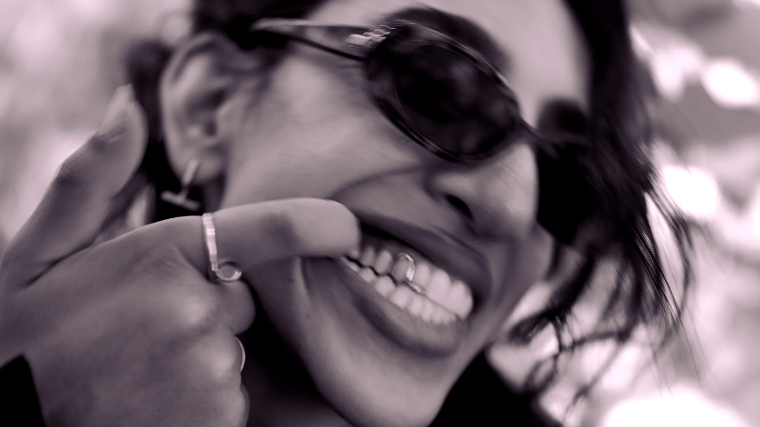 Close-up of a person with dark hair and sunglasses, showing a big smile, holding a phone near their face.