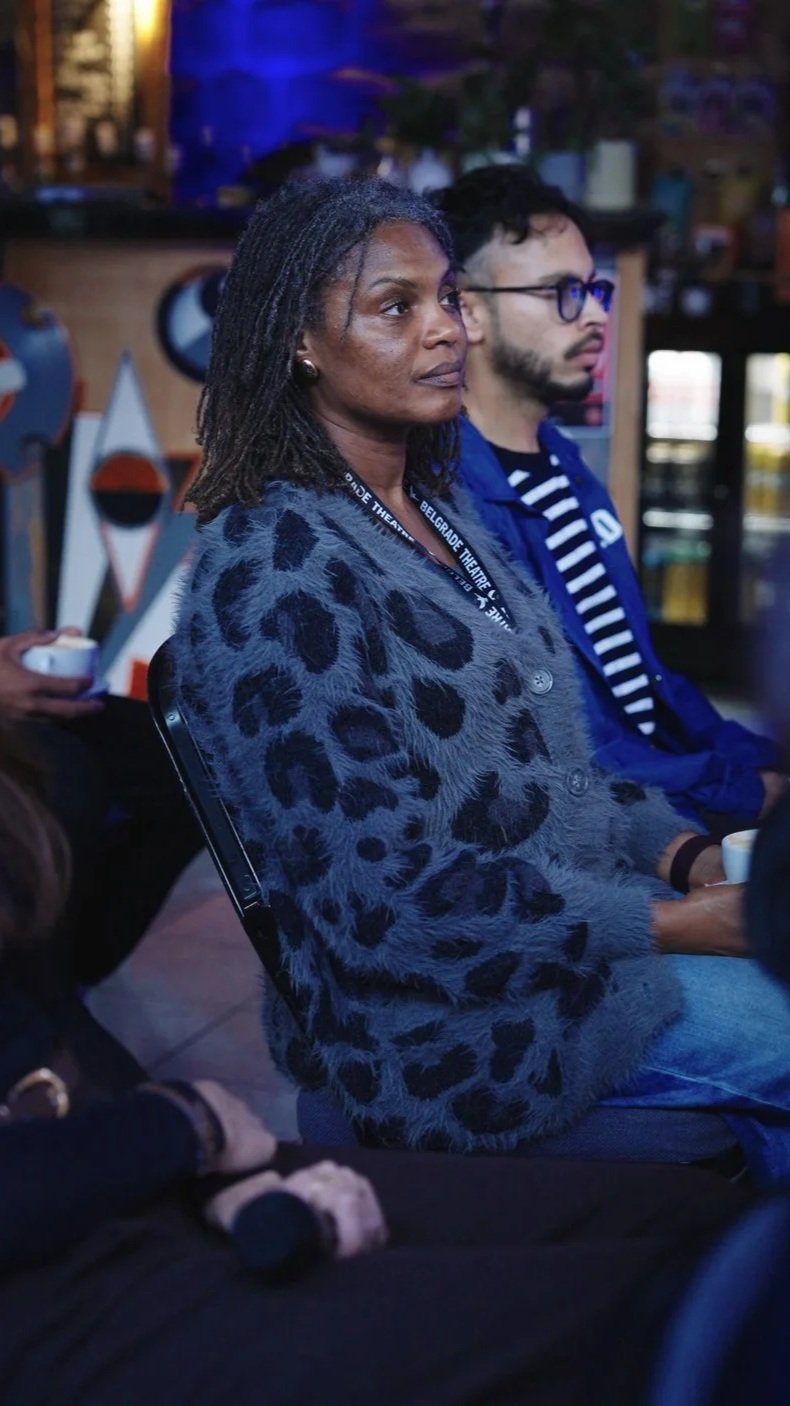 Two people sitting at a table in a dimly lit venue, with a woman in the foreground with dreadlocks, wearing a leopard print sweater, and a man in the background with glasses and a striped shirt.