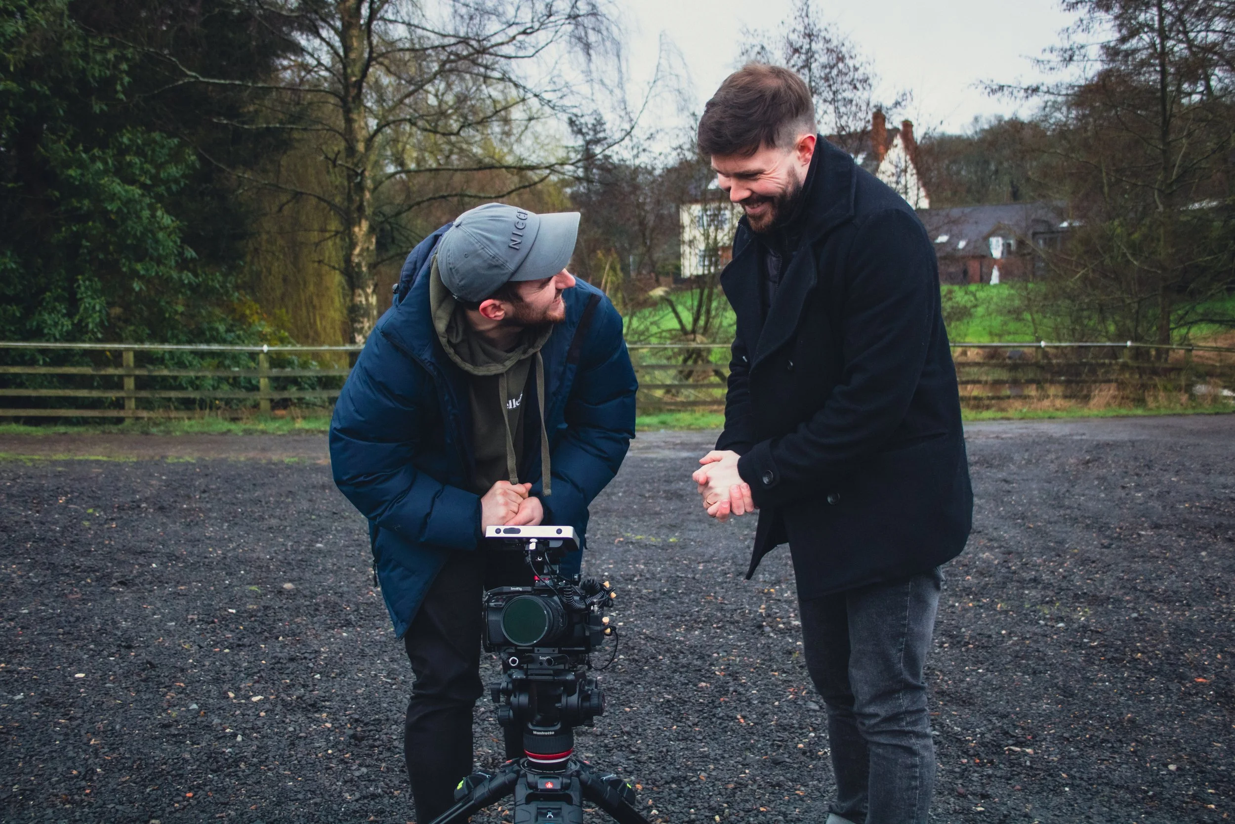 Two men are outdoors, standing on a gravel path, engaging in a joyful conversation. One man holds a camera mounted on a tripod, and they are smiling at each other. The background features trees and a few houses under a cloudy sky.