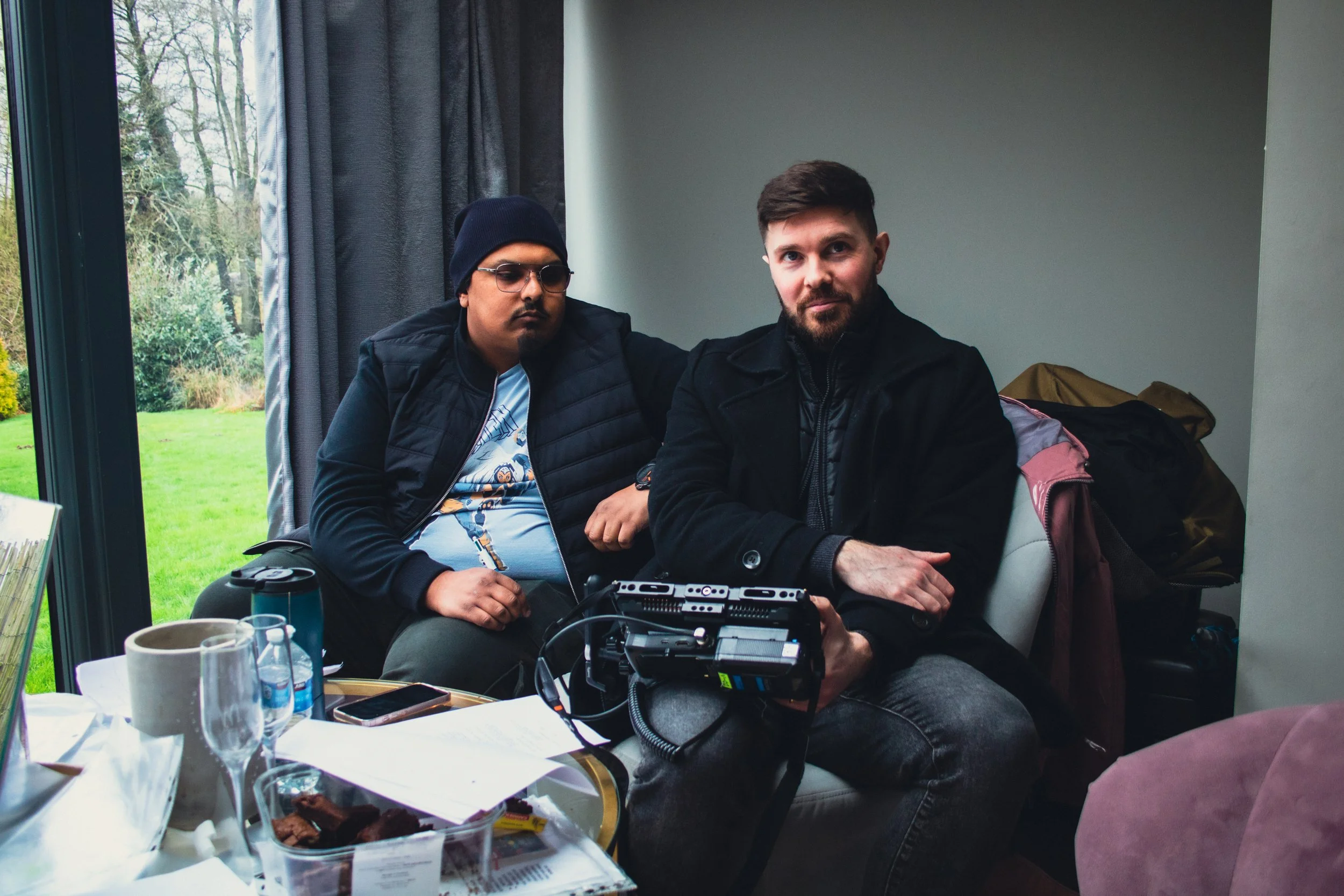 Two men sitting indoors at a table with food, drinks, and electronic equipment, with large window and greenery outside.
