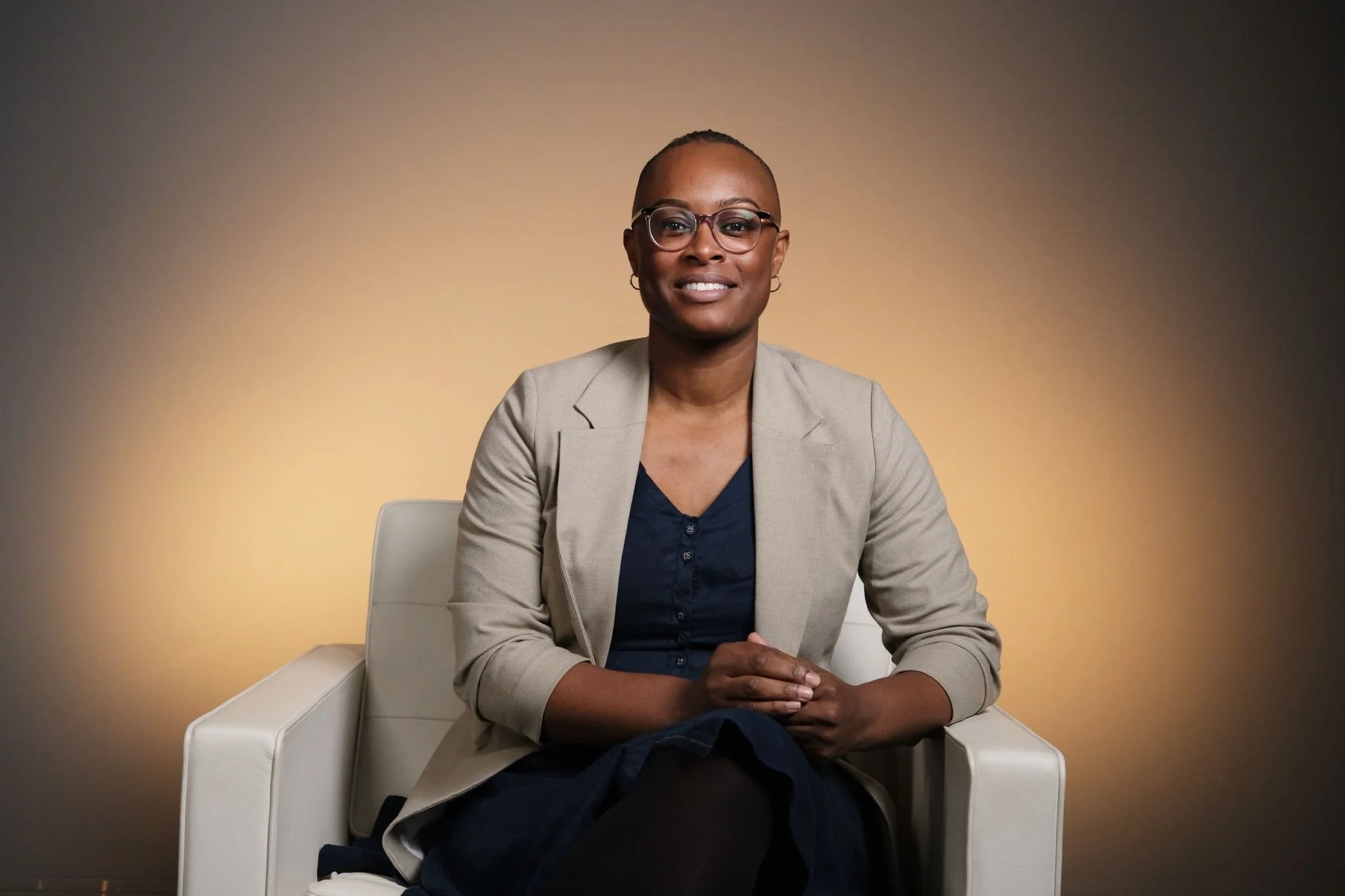 A woman with a shaved head, glasses, and earrings sitting in a white armchair against a plain gradient background. She is wearing a beige blazer over a dark top and has her hands folded on her lap, smiling slightly at the camera.