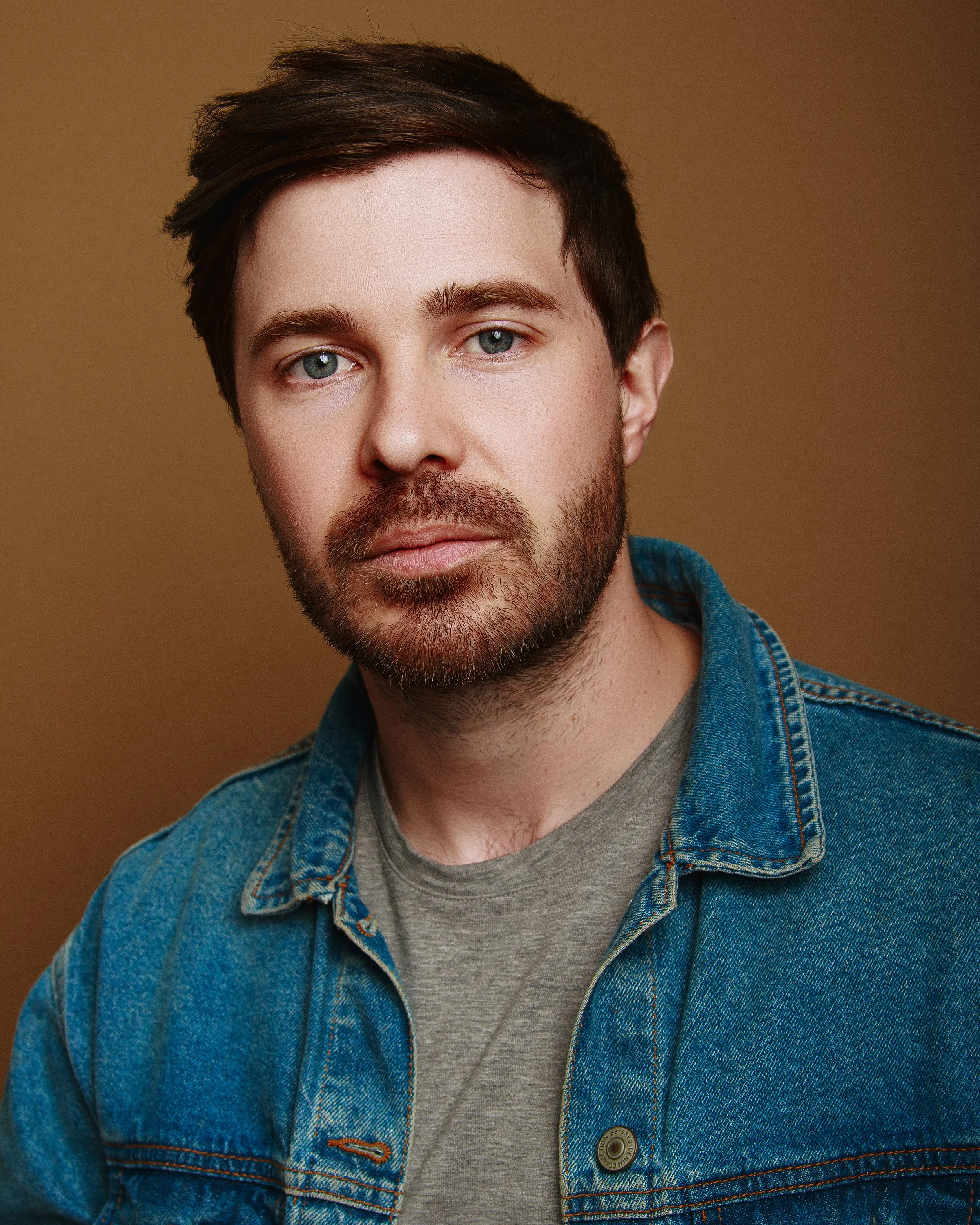 Close-up of a young man's face with blue eyes and dark brown hair, wearing a denim jacket over a gray t-shirt, against a brown background.