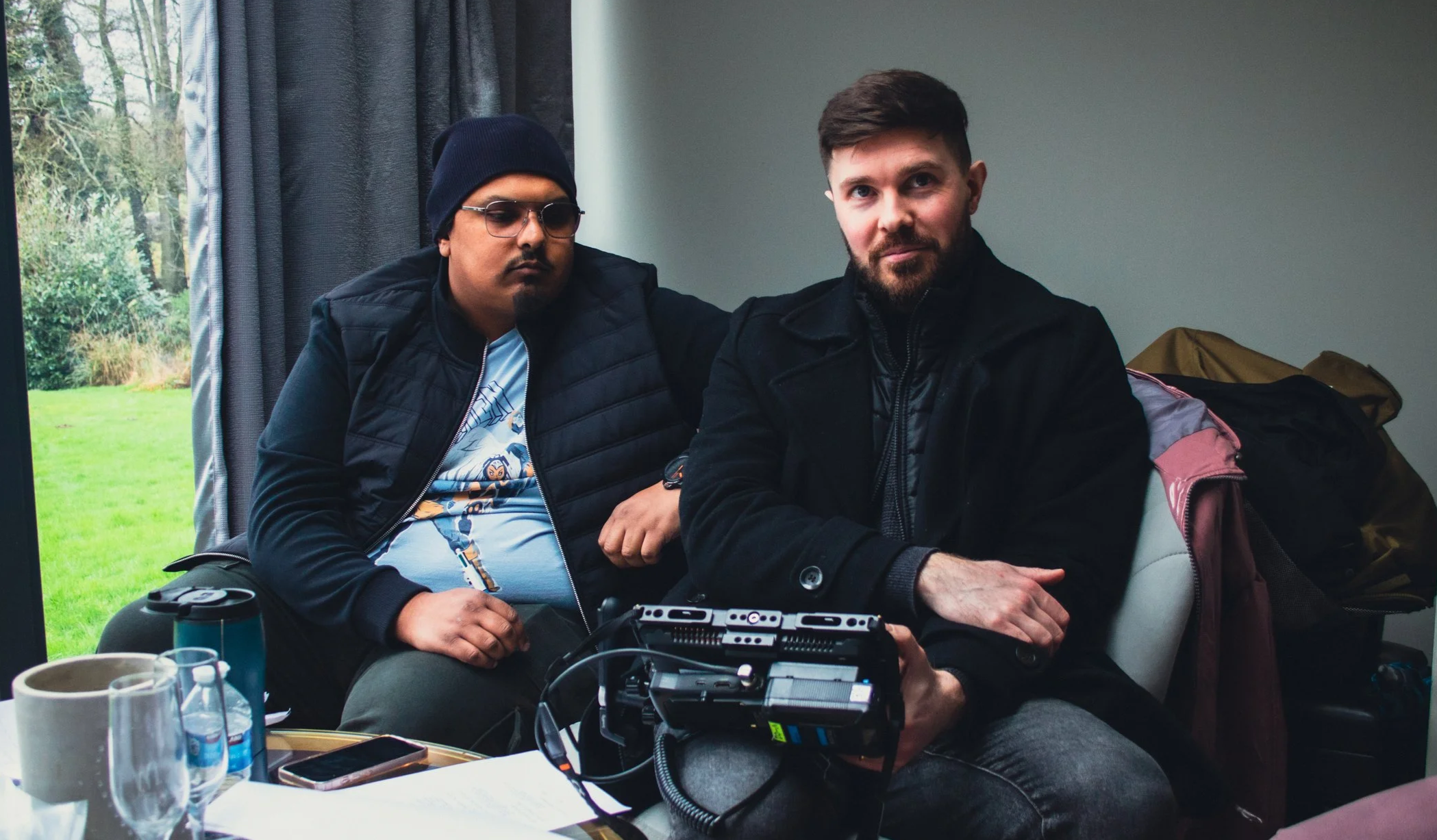 Two men sitting indoors, with a table that has bottles and glasses, a camera or audio equipment, and papers. One man is wearing glasses and a beanie, the other has a beard and short hair. There is a window with curtains and trees outside in the background.