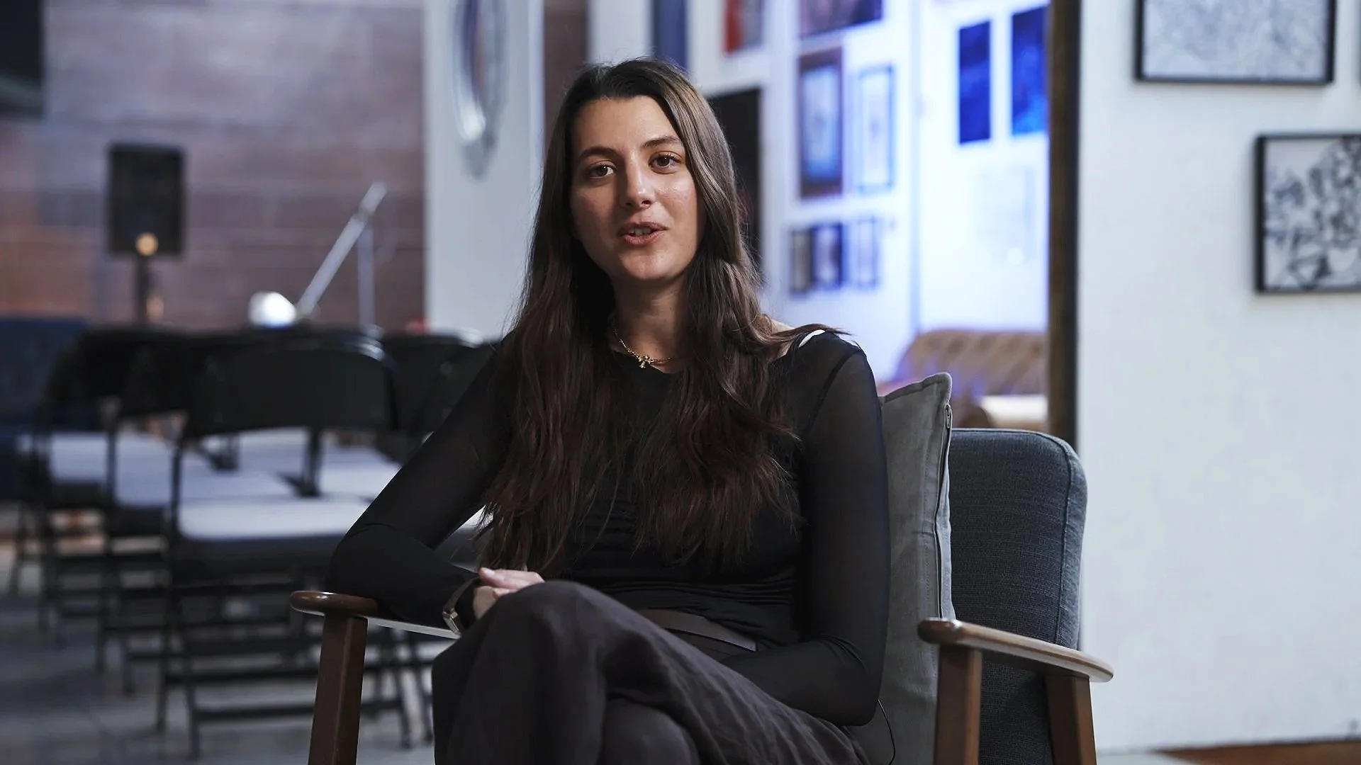 A woman with long dark hair, wearing a black top, sitting on a gray armchair in a modern interior space decorated with framed pictures on the wall.