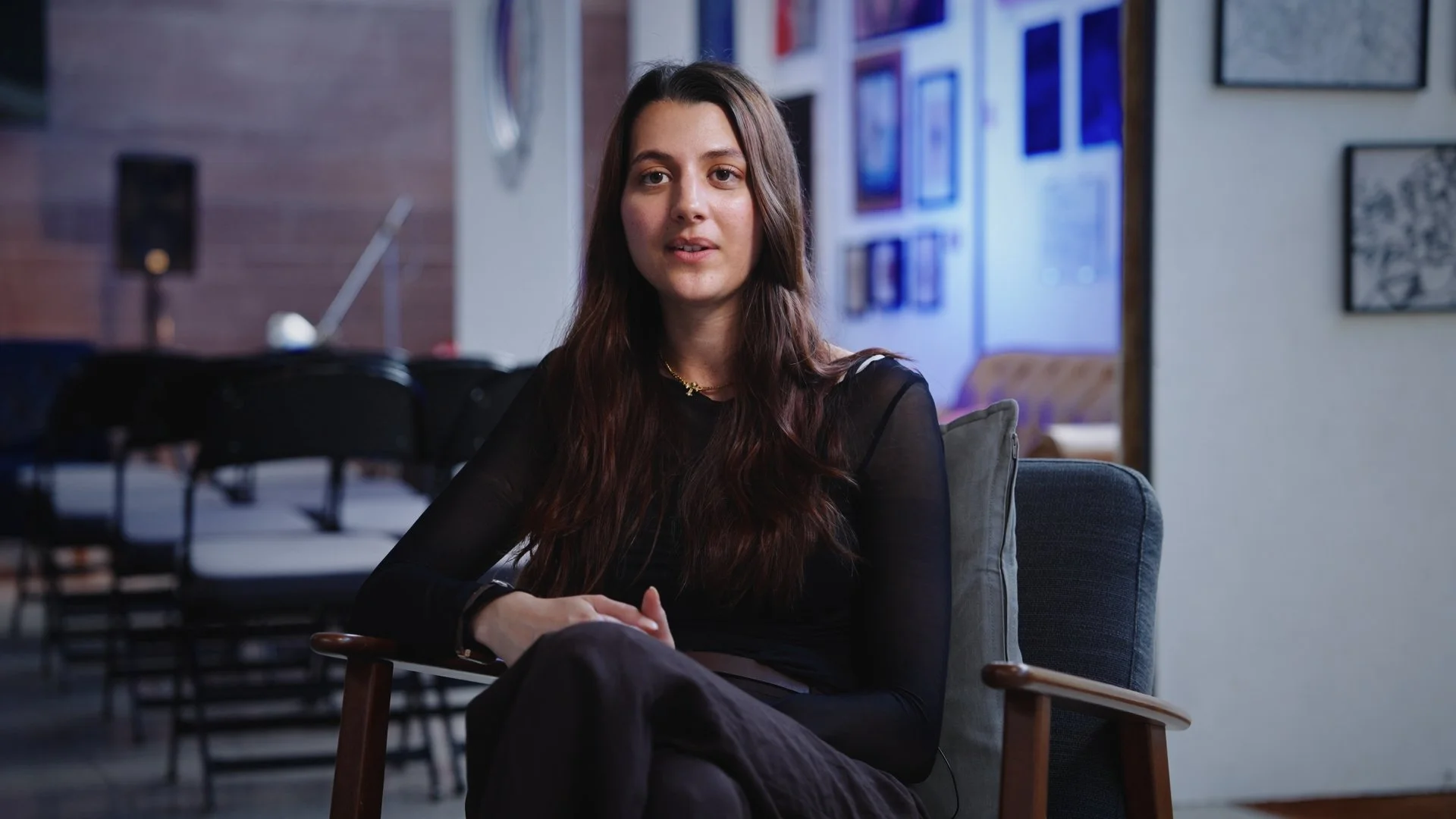 A young woman with long brown hair sitting on a chair in a room with framed pictures on the wall, looking slightly to the side.