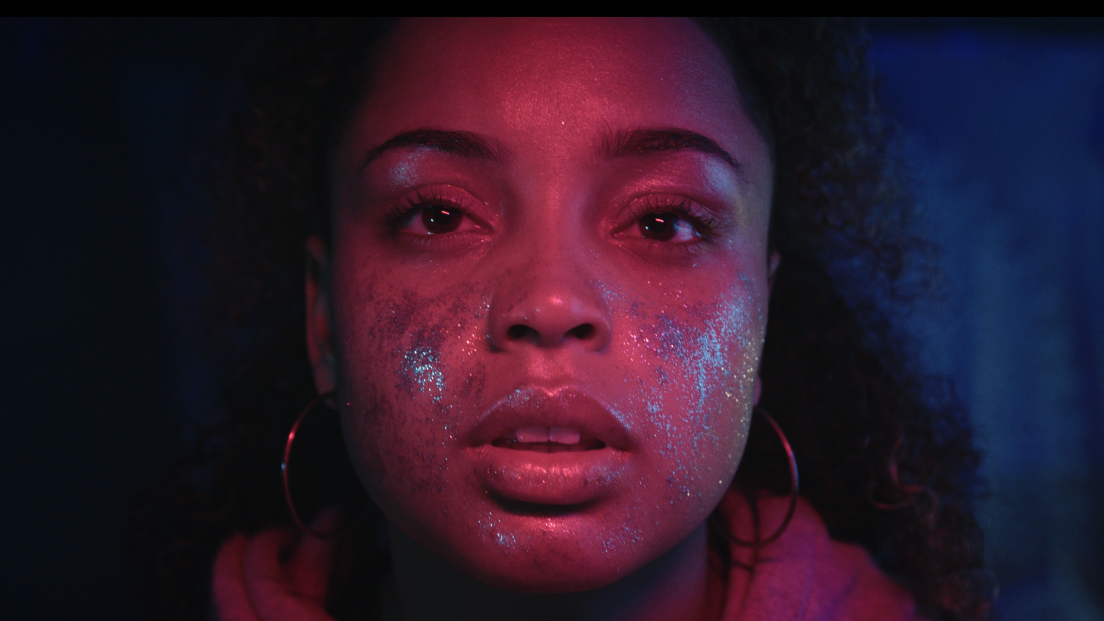 A close-up portrait of a woman with dark curly hair, neon pink and blue lighting, and sparkles on her face.
