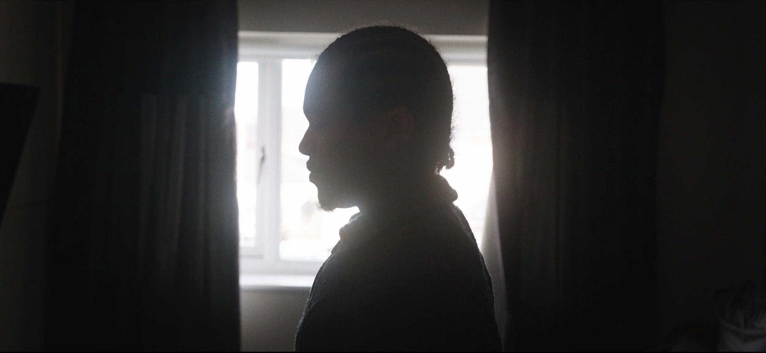 A person with curly hair is standing indoors near a window with light streaming in, creating a silhouette.