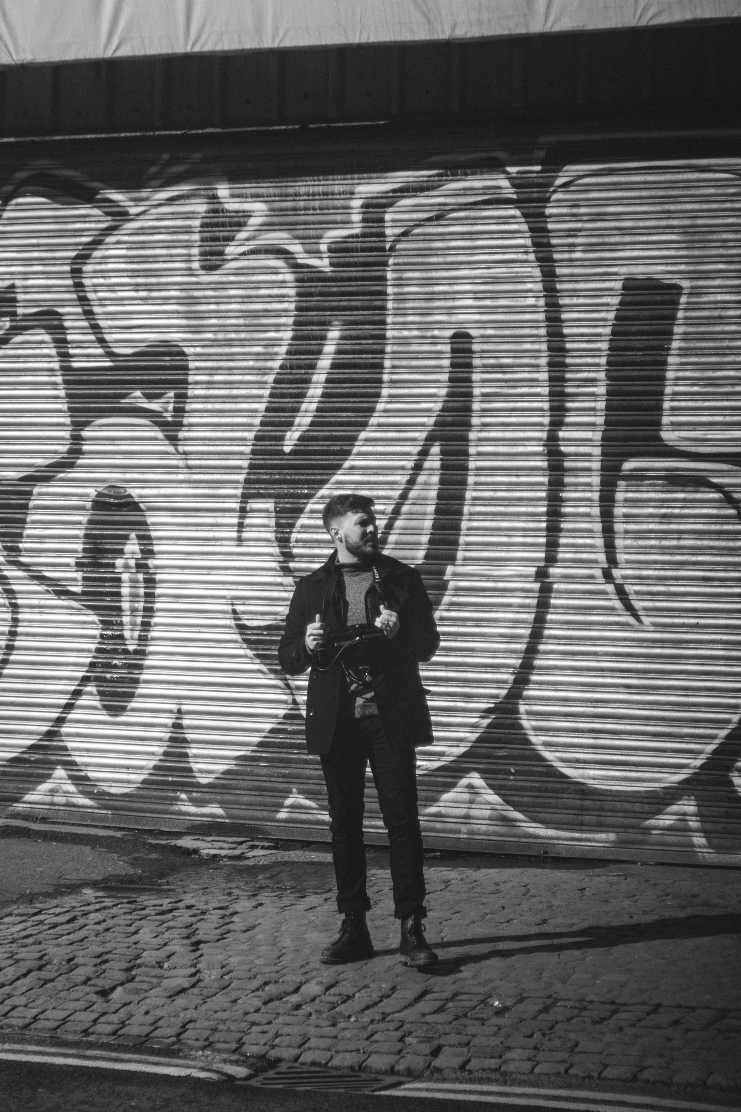 A man with a beard and short hair wearing a black jacket, holding a camera, standing in front of a large graffiti mural on a metal shutter door. The graffiti is in bold, stylized letters.