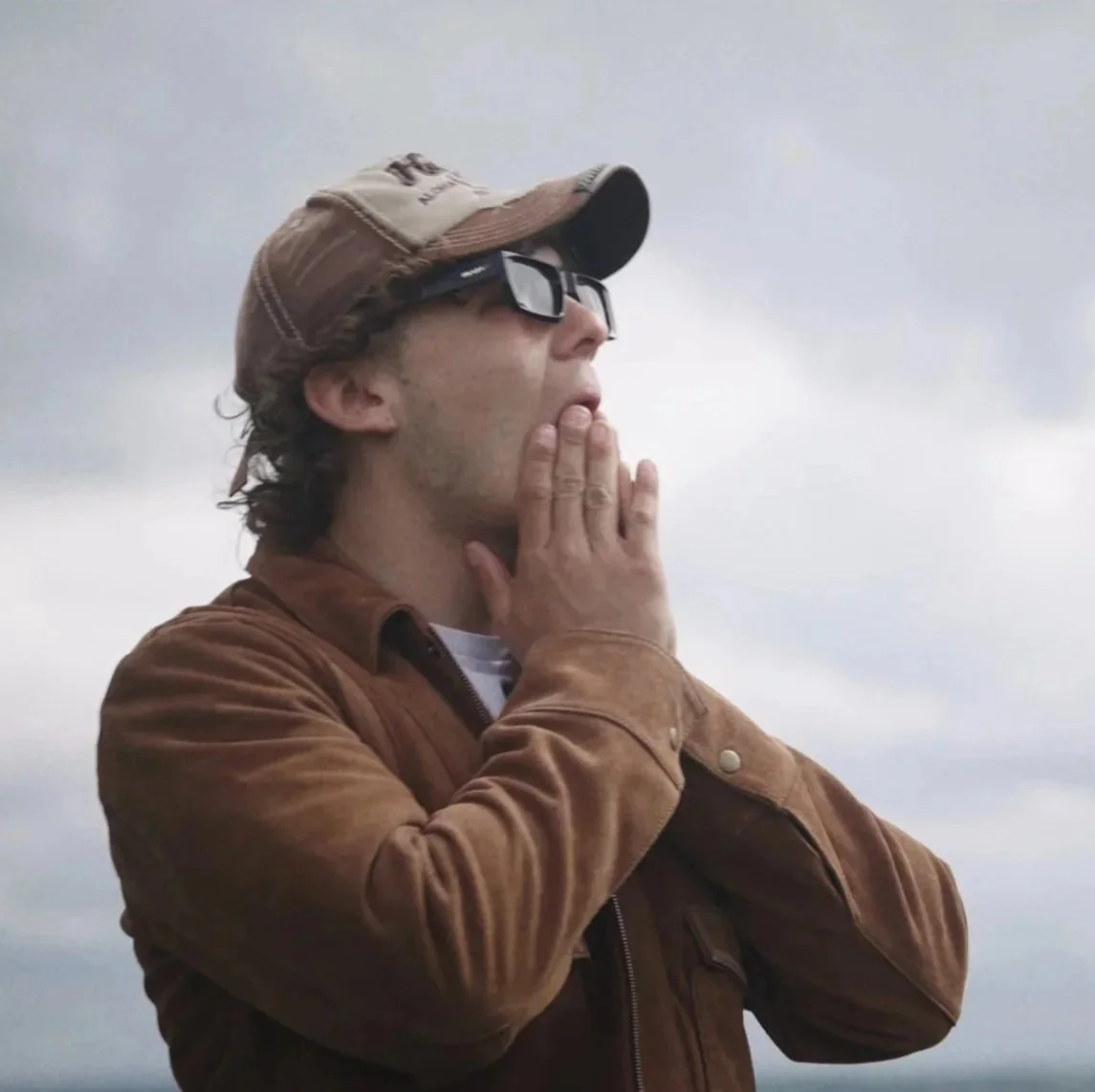 A man wearing a brown cap, sunglasses, and a brown jacket, with his hand near his mouth, looking at the sky against a cloudy sky background.
