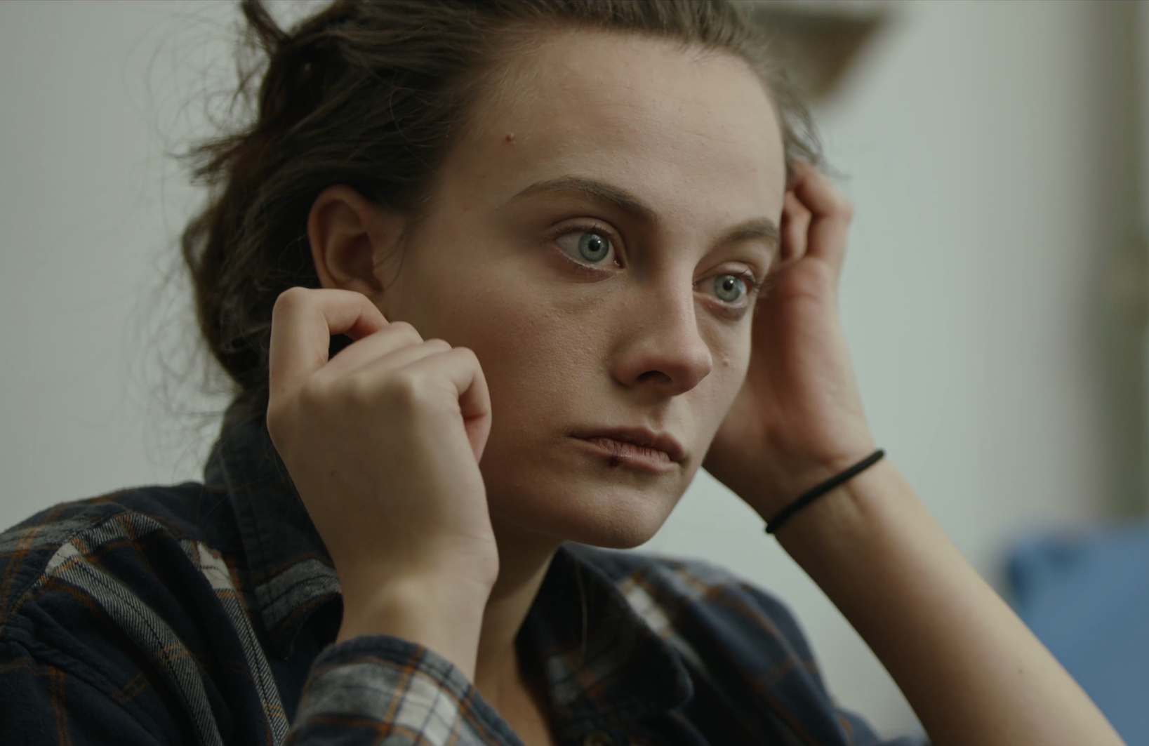 A young woman with green eyes, brown hair, and fair skin is seen adjusting her hair while looking pensively.