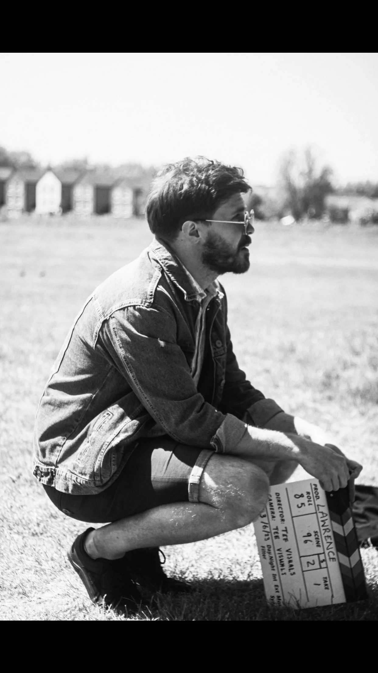 Black and white photo of a man sitting outdoors on grass, holding a clapperboard, wearing sunglasses and a denim jacket, with blurred residential buildings in the background.