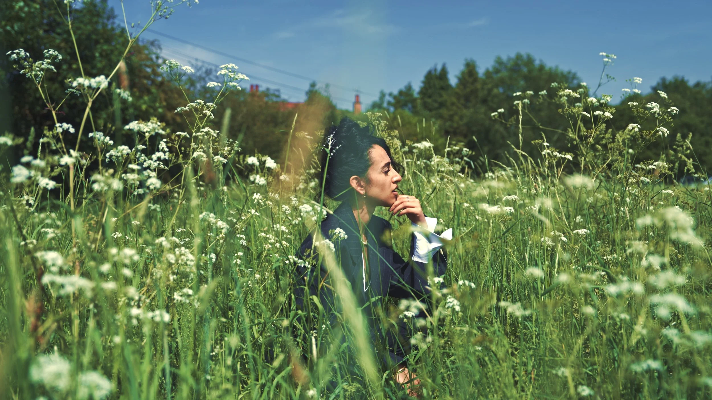 Woman in formal attire, wearing white gloves, sitting in a field of white wildflowers, with her eyes closed and hand near her face on a sunny day.
