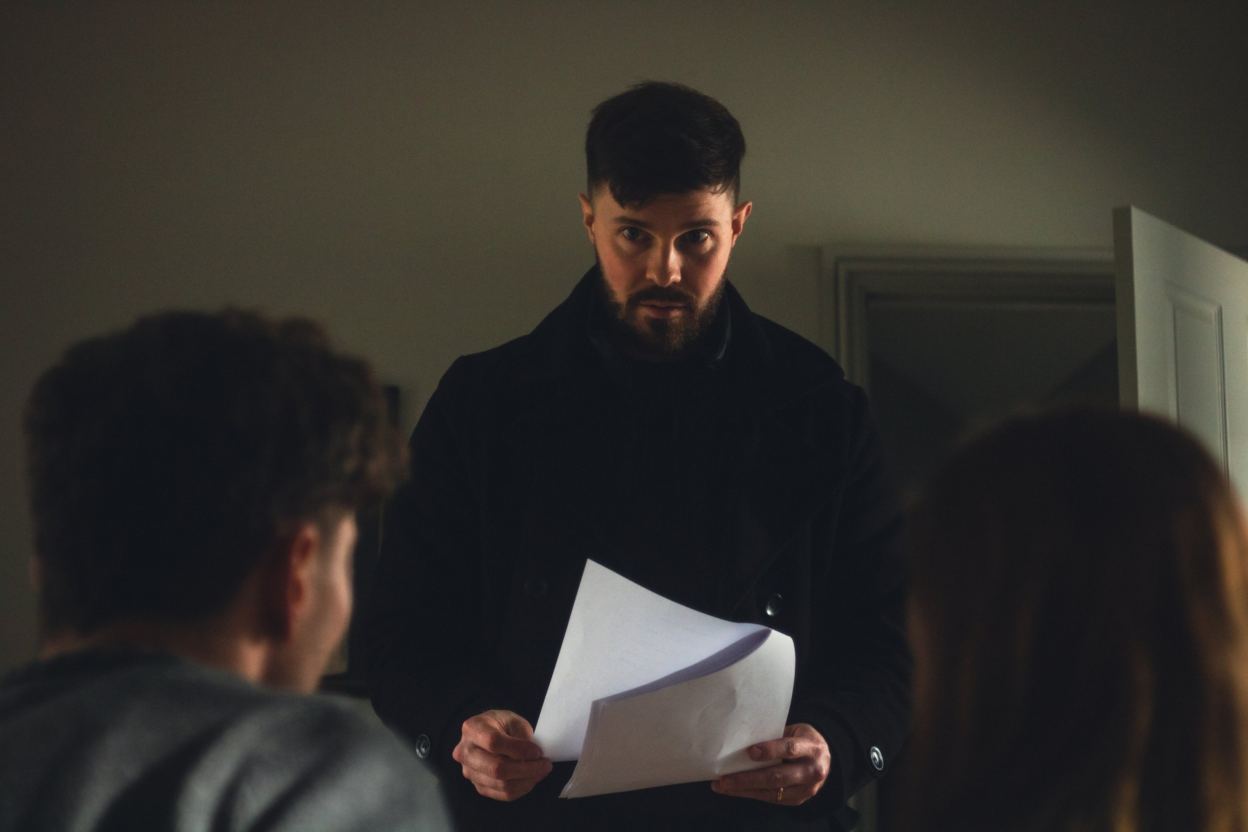 A man with dark hair and a beard wearing a dark shirt, looking directly at the camera while holding papers, with two blurred individuals in the foreground and a partially open door in the background.