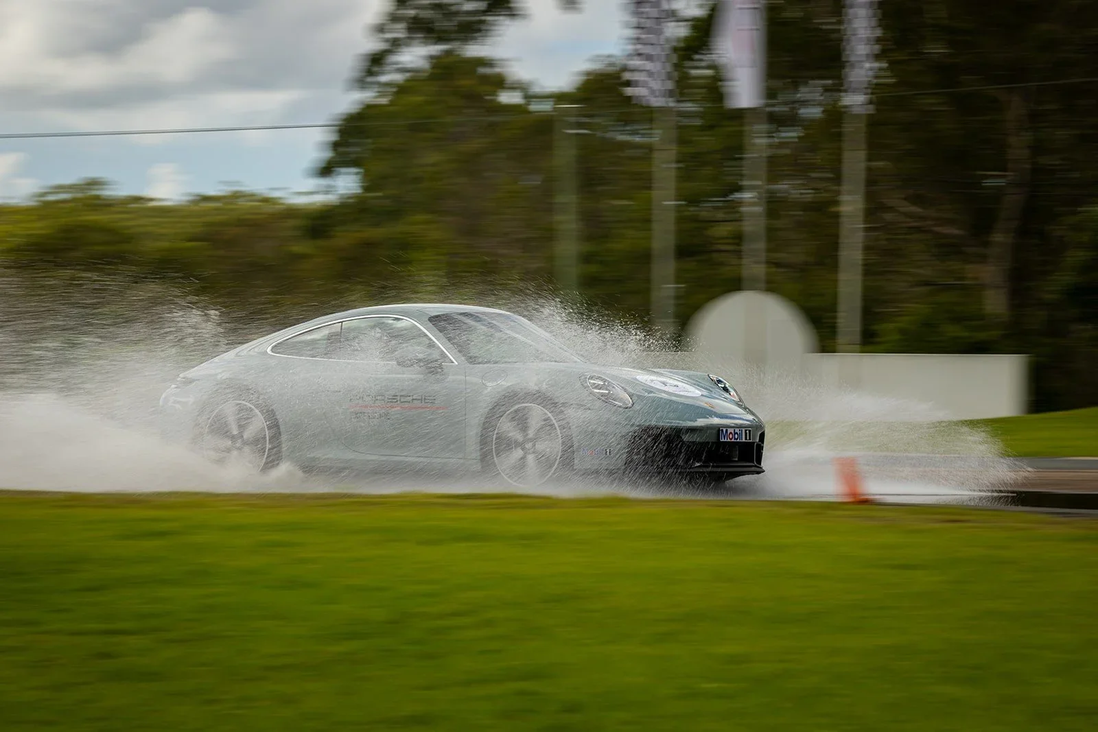 A silver Porsche racing car speeding through a wet track, creating a spray of water around it, with trees and poles in the background.