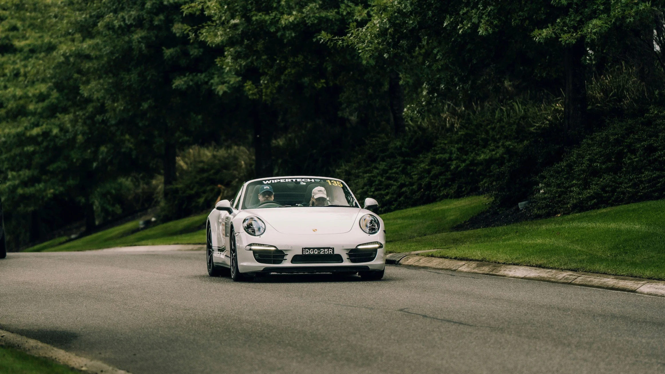 A white Porsche sports car driving on a curved road surrounded by green trees and grass.
