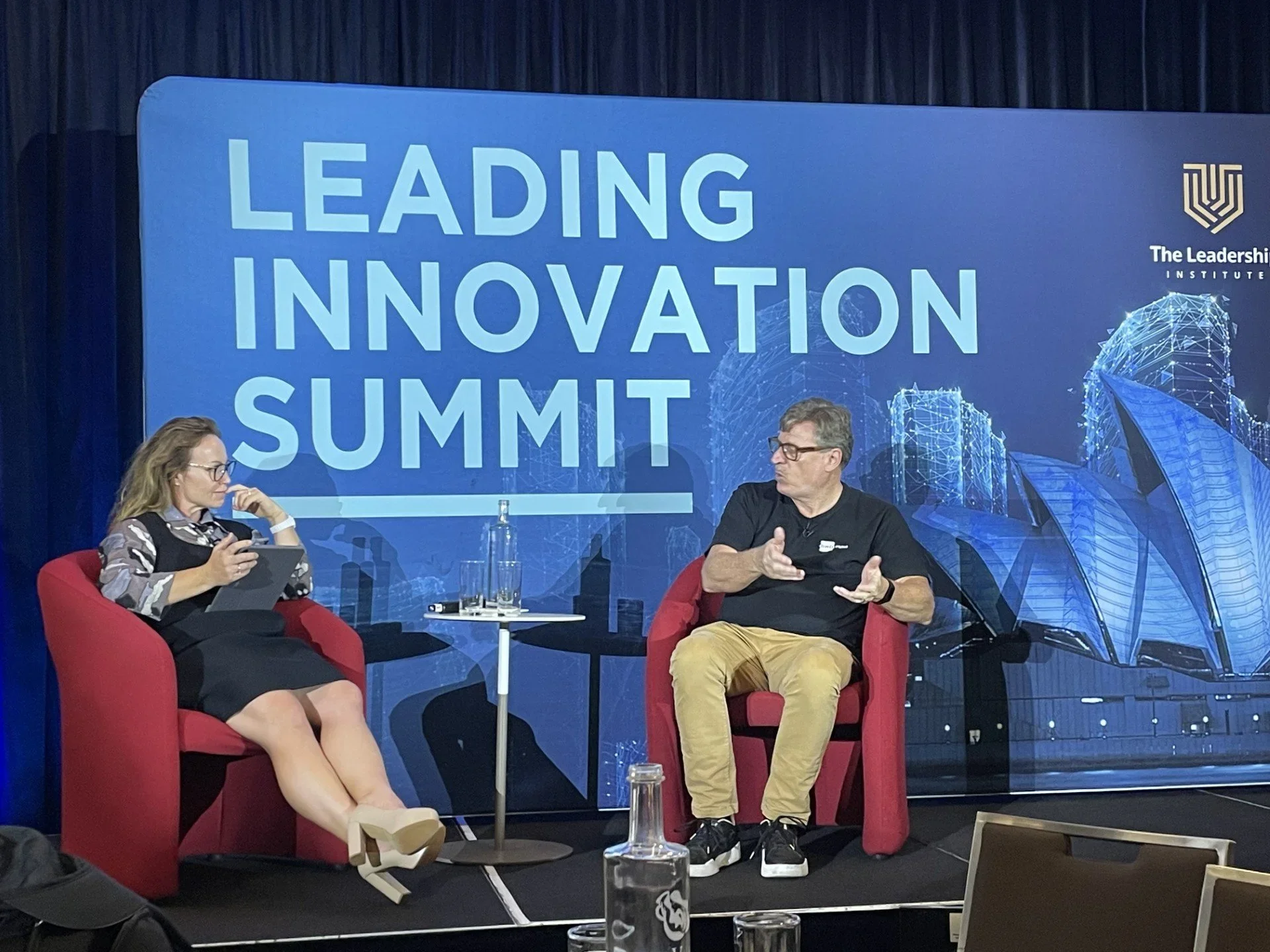 Two people sit on red chairs with a small white side table between them, participating in a panel discussion at the Leadership Innovation Summit. The backdrop features blue graphics and large text reading 'Leading Innovation Summit,' along with the logo for The Leadership Institute.