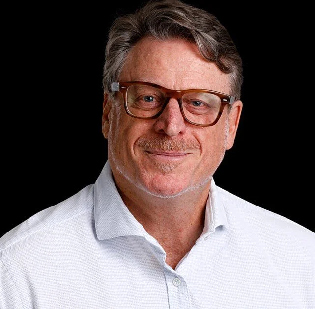 Portrait of a middle-aged man with gray hair, glasses, and a slight smile, wearing a white collared shirt against a black background.