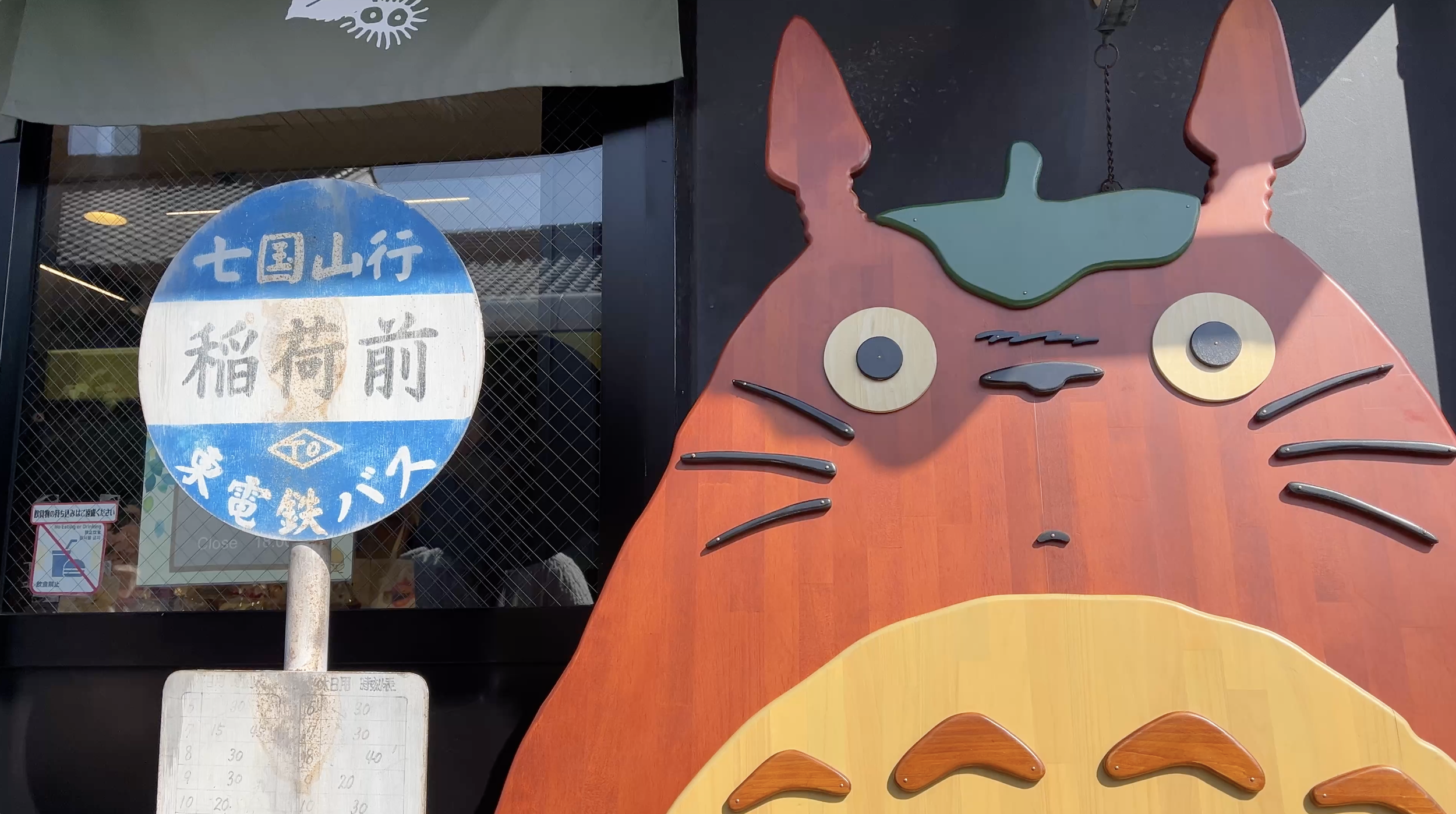 A storefront sign with Japanese characters and a cartoonish face resembling a cat or animal, with a large head, big eyes, and pink ears, set against a black background. There is also a round blue and white sign with Japanese text.