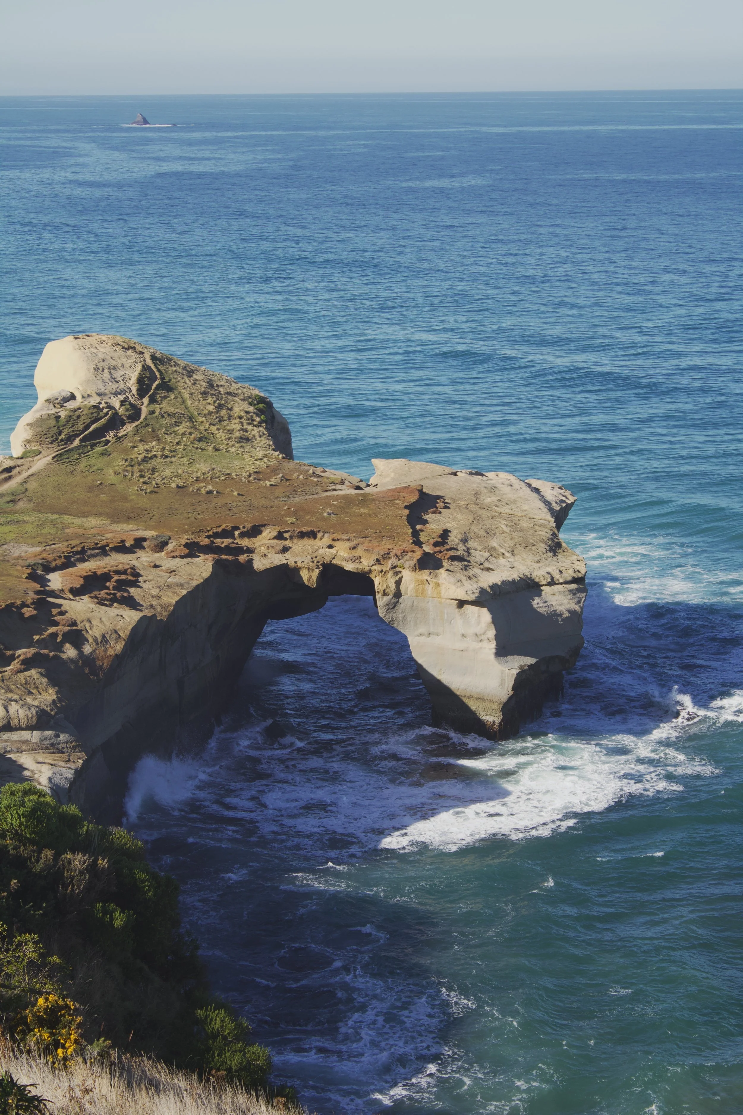 Ocean waves crashing against rocky shoreline with natural arches and green vegetation in foreground, distant islands on horizon under clear sky.