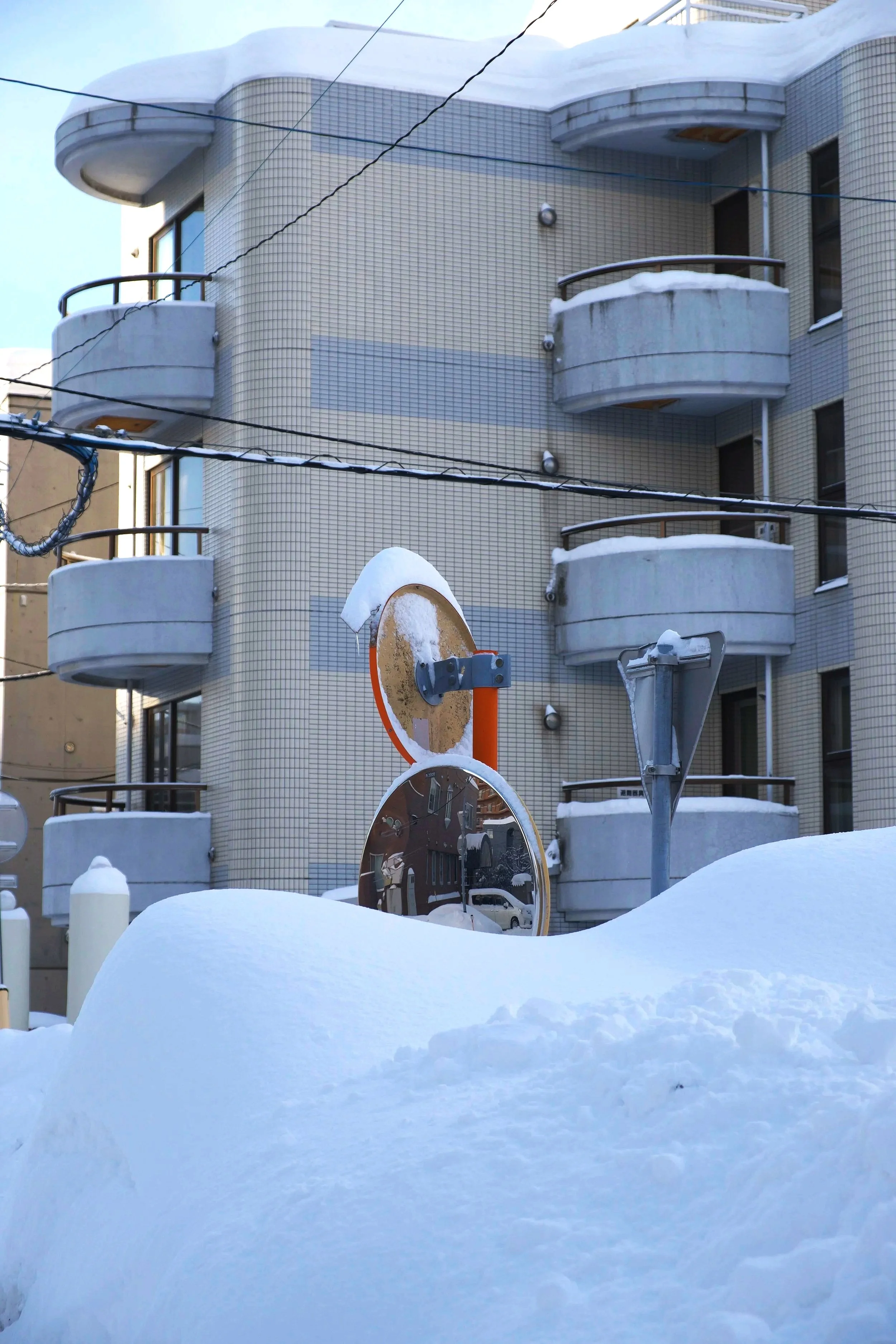 A building with balconies covered in snow, utility wires crisscrossing the scene, and a snow-covered street with a reflective traffic mirror in the foreground.