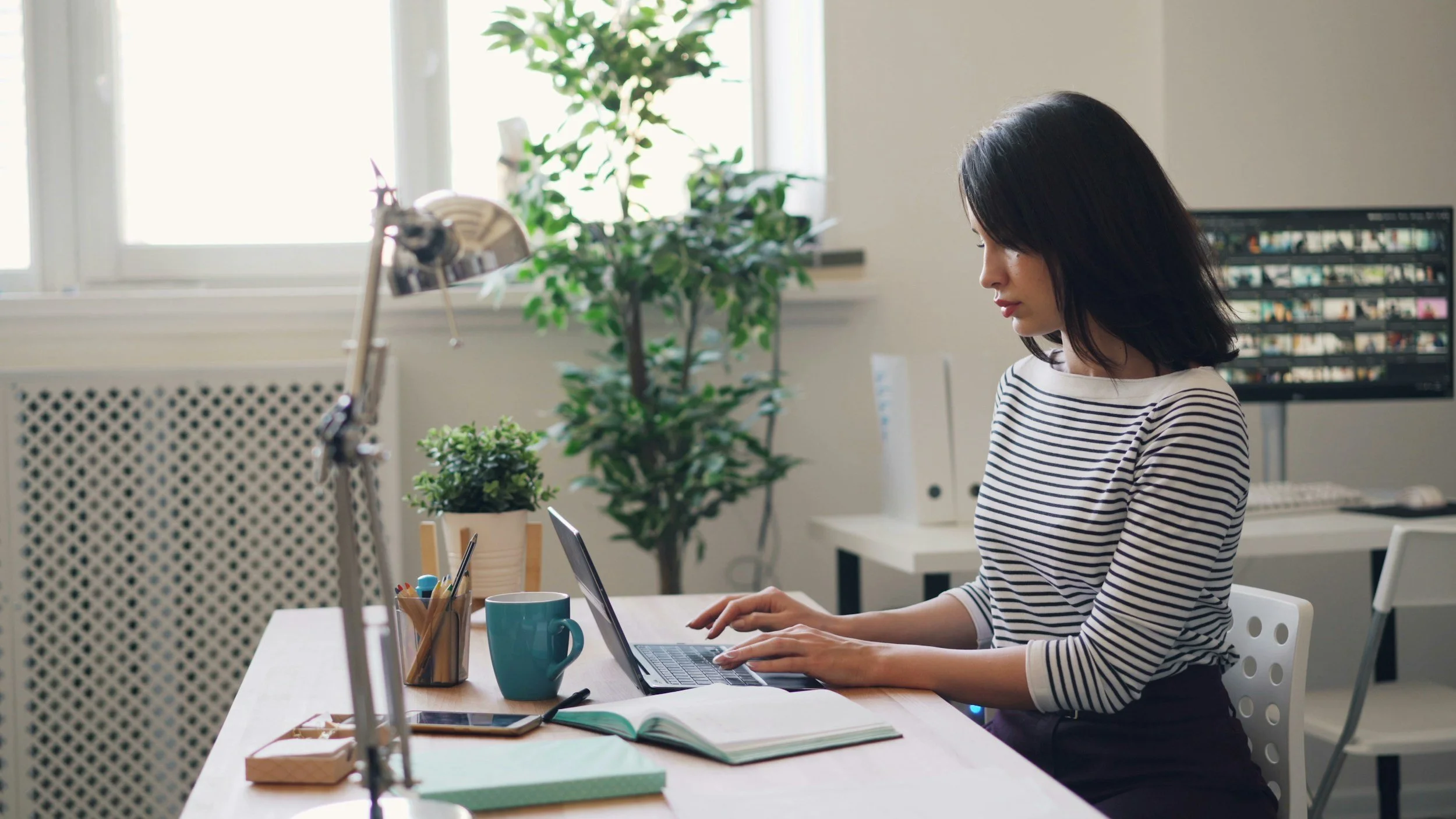 Woman with black hair wearing a striped shirt working on a laptop at a desk with a green mug, notebooks, and pens, in a bright office with plants and a computer monitor in the background.