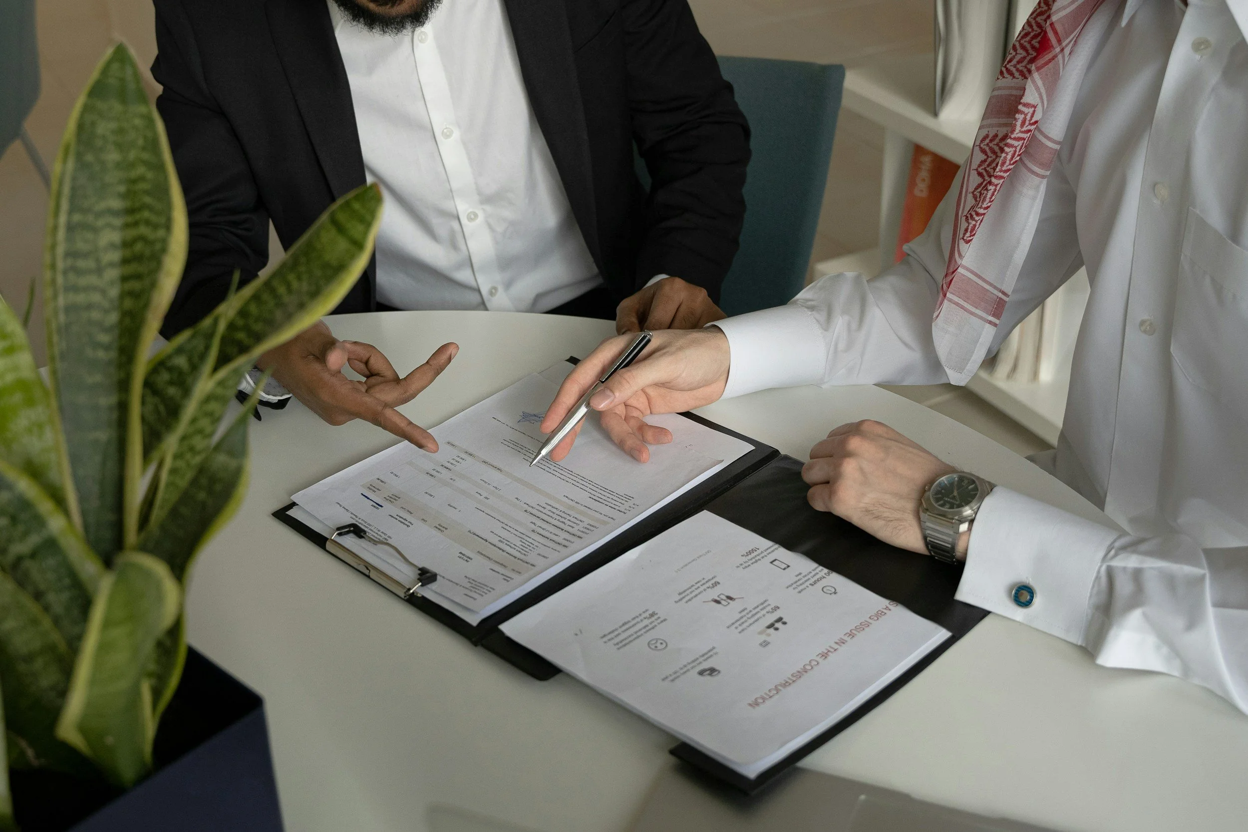 Two men in professional attire reviewing documents and signing papers on a white table, with a potted plant partially visible in the foreground.