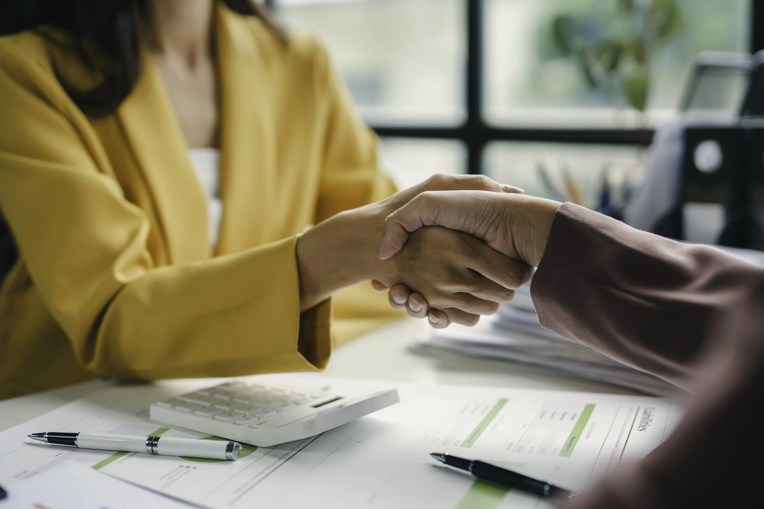 Two people shaking hands over a desk with papers, pens, and a calculator, indicating a business or work meeting in an office setting.