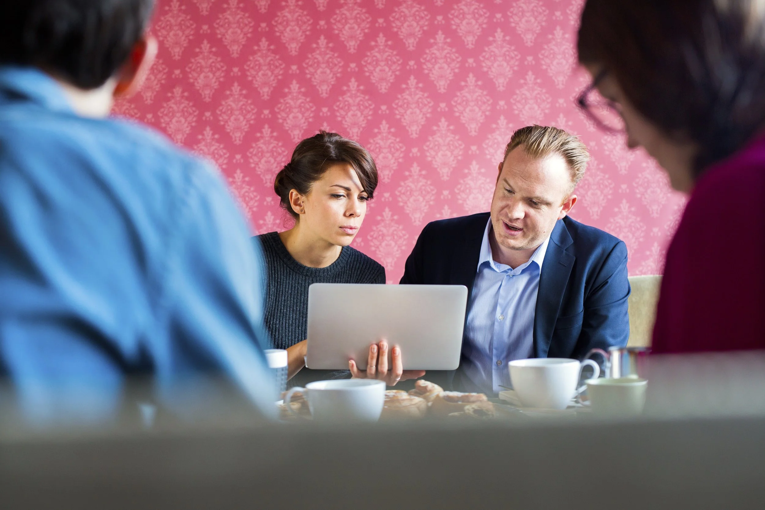 Four people sitting at a table, looking at a laptop, with a pink wallpaper background.