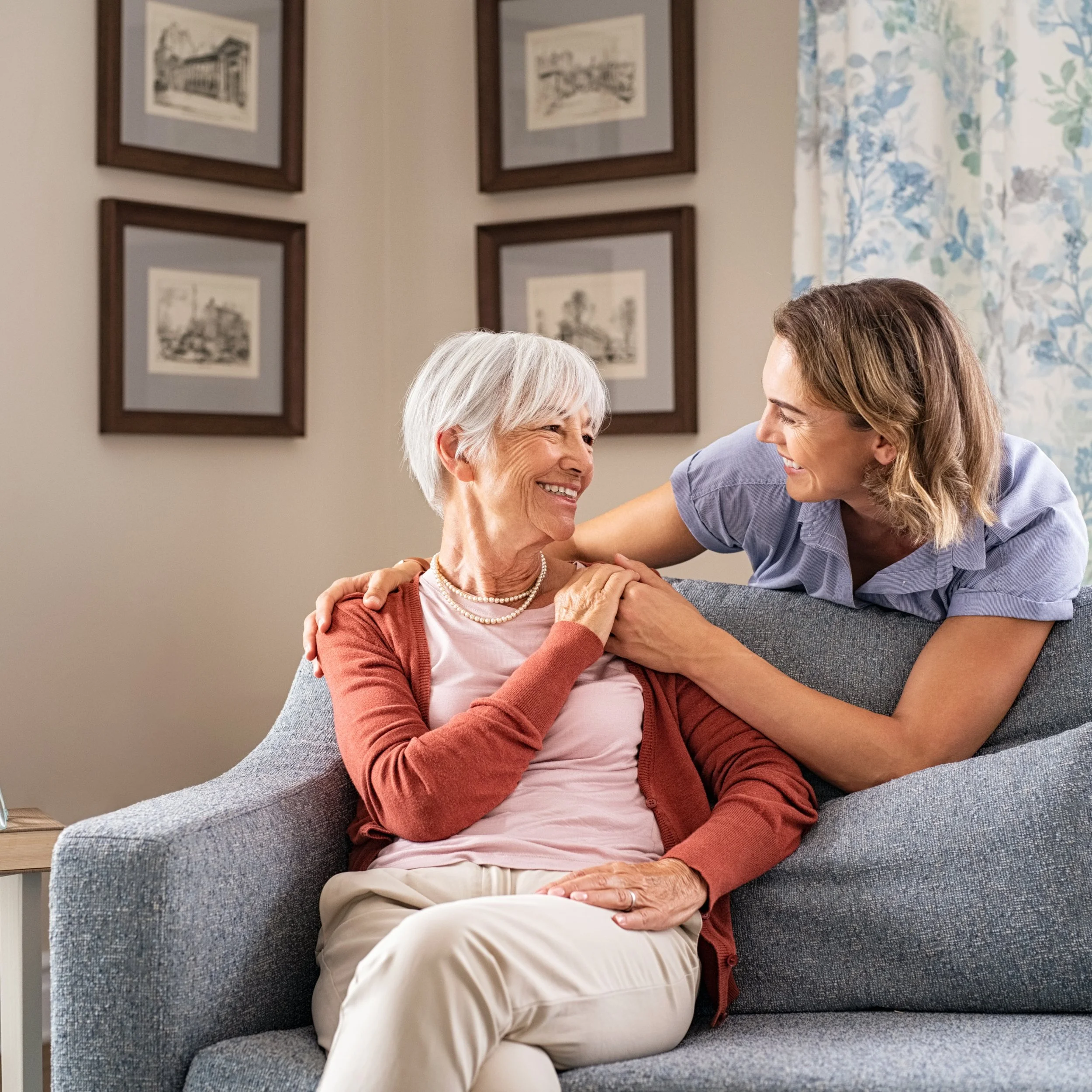 A young woman and an elderly woman are smiling and embracing on a gray couch in a living room with framed artwork and floral curtains in the background.