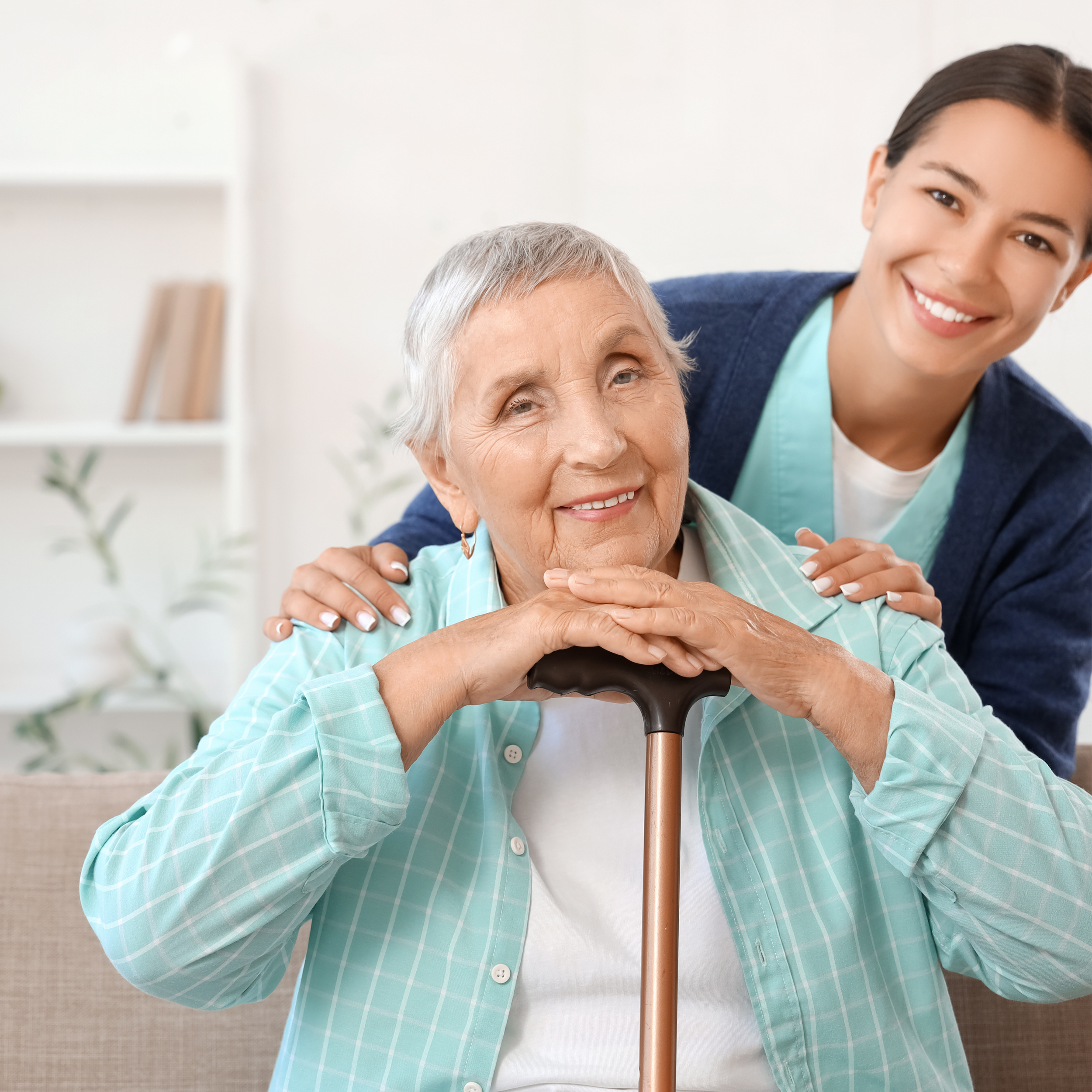 A young woman with dark hair smiling and leaning close to an elderly woman with gray hair, who is holding a cane, in a bright room with white walls and shelves.