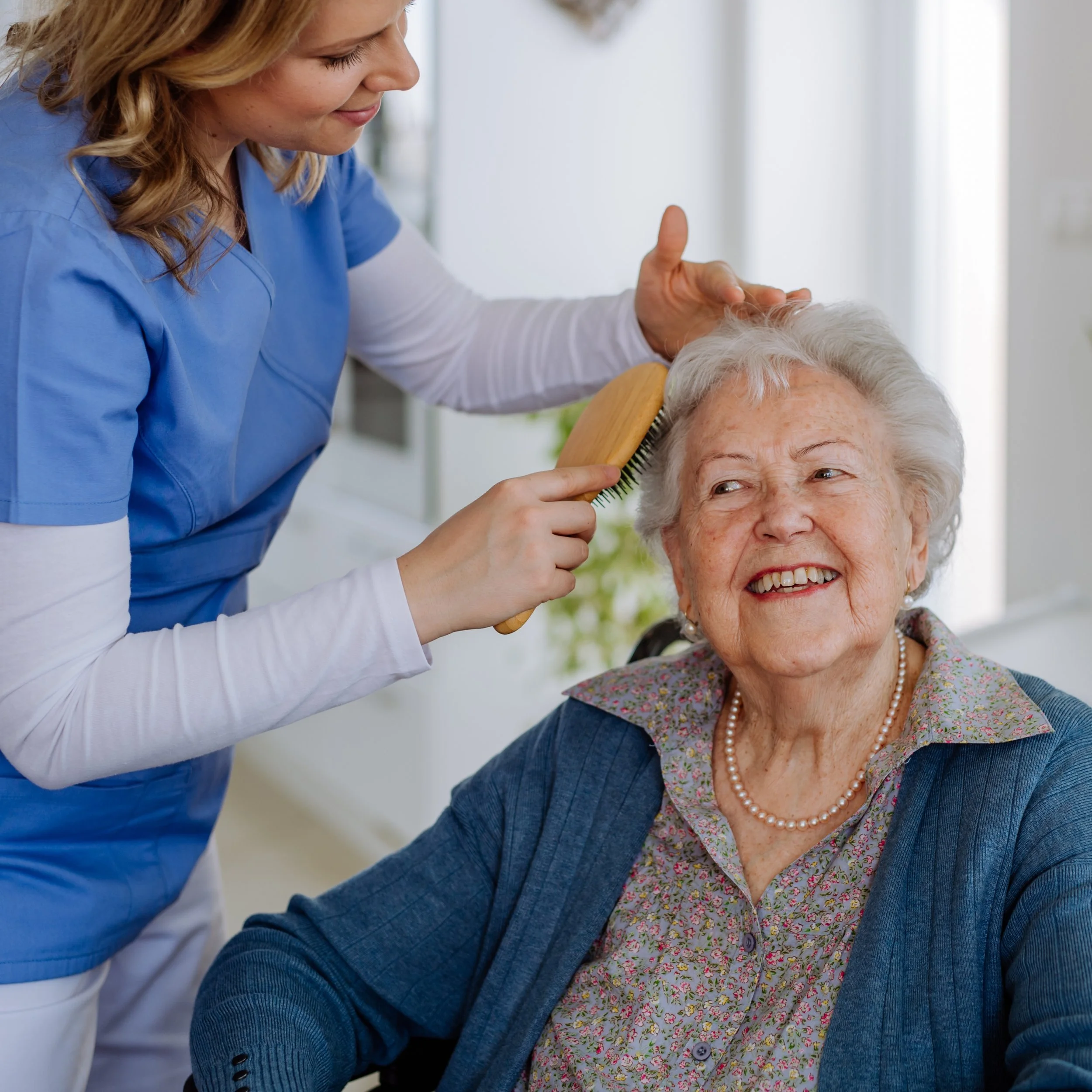 A caregiver styling an elderly woman's white hair with a hairbrush in a bright, cheerful room.