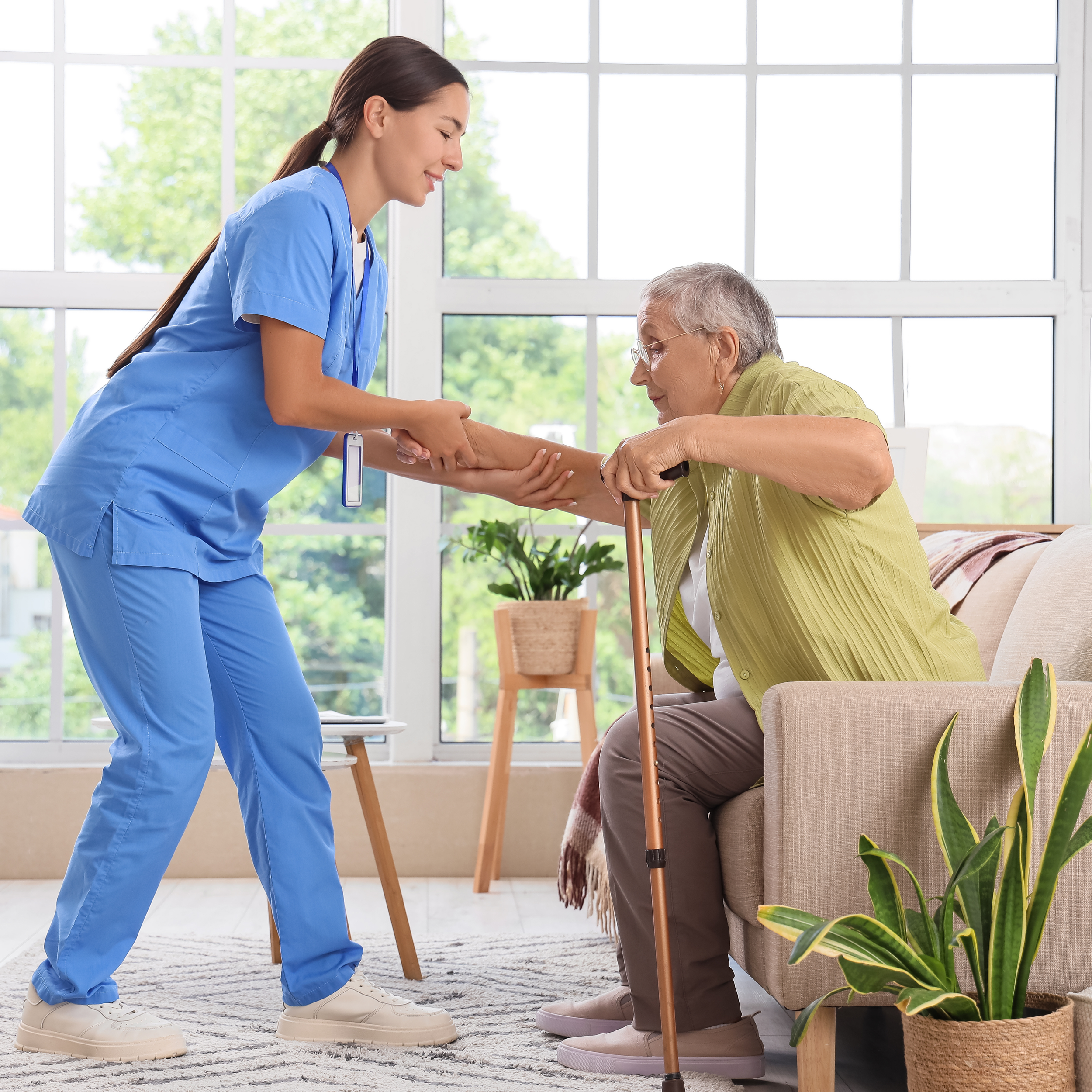 A female nurse in blue scrubs assisting an elderly woman with her arm in a well-lit room with large windows, green trees outside, and indoor plants.