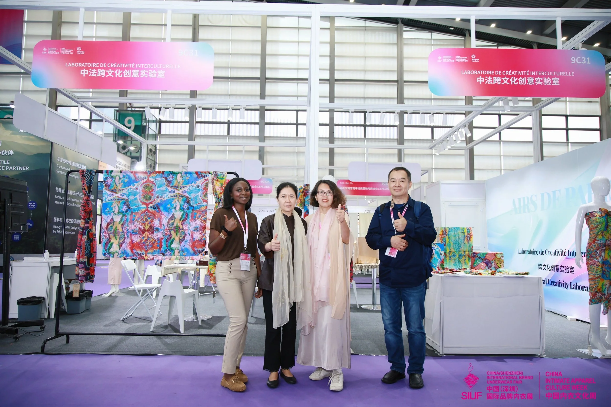 Groupe de quatre personnes debout devant un stand d'exposition avec des tissus colorés, à l'intérieur d'un bâtiment avec une structure en métal et des panneaux en verre. Le stand porte un panneau indiquant un laboratoire de créativité interculturelle.