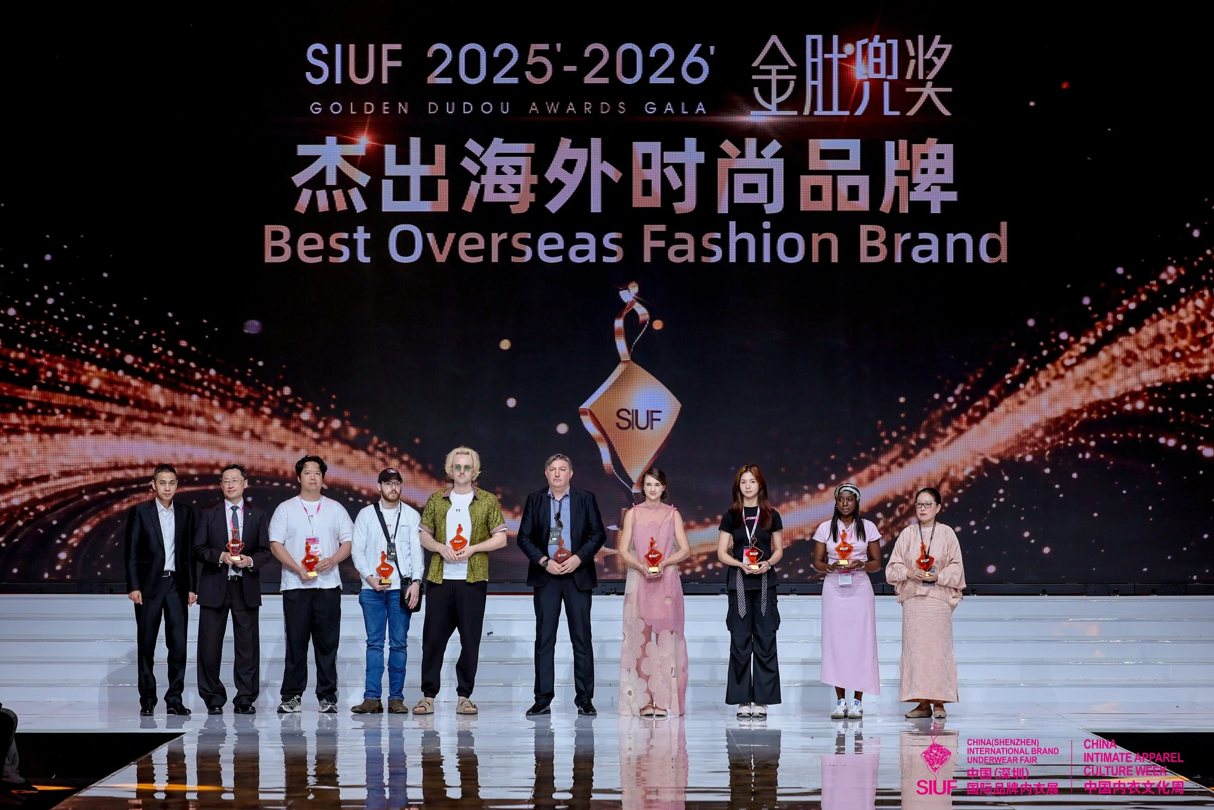 Groupe de personnes debout sur scène recevant des trophées lors d'une cérémonie, avec un grand écran en arrière-plan affichant le texte 'SIUF 2025-2026', 'Golden Dudou Awards Gala', et 'Best Overseas Fashion Brand'.