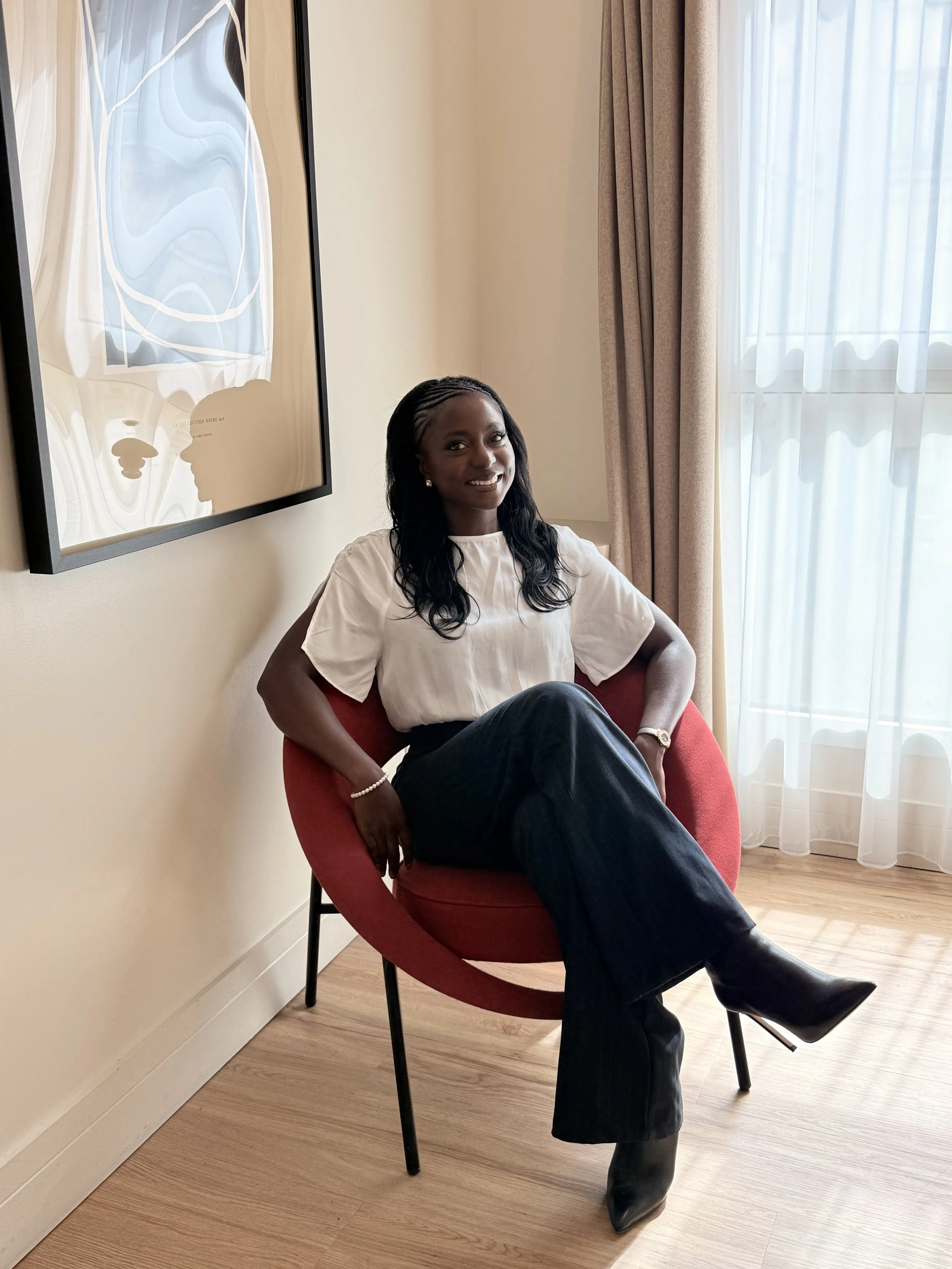Une femme assise dans un fauteuil rouge, souriant, dans une pièce lumineuse avec une fenêtre à rideaux blancs. Elle porte un pantalon noir, une chemise blanche et des bottes noires.