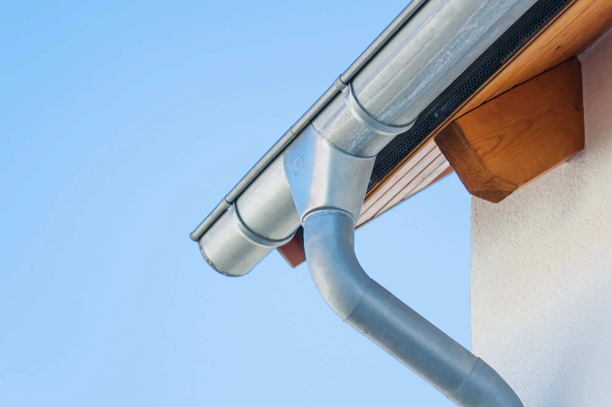 Close-up of a metal gutter and downspout attached to the corner of a building with a wooden eaves and a textured white wall, against a bright blue sky.