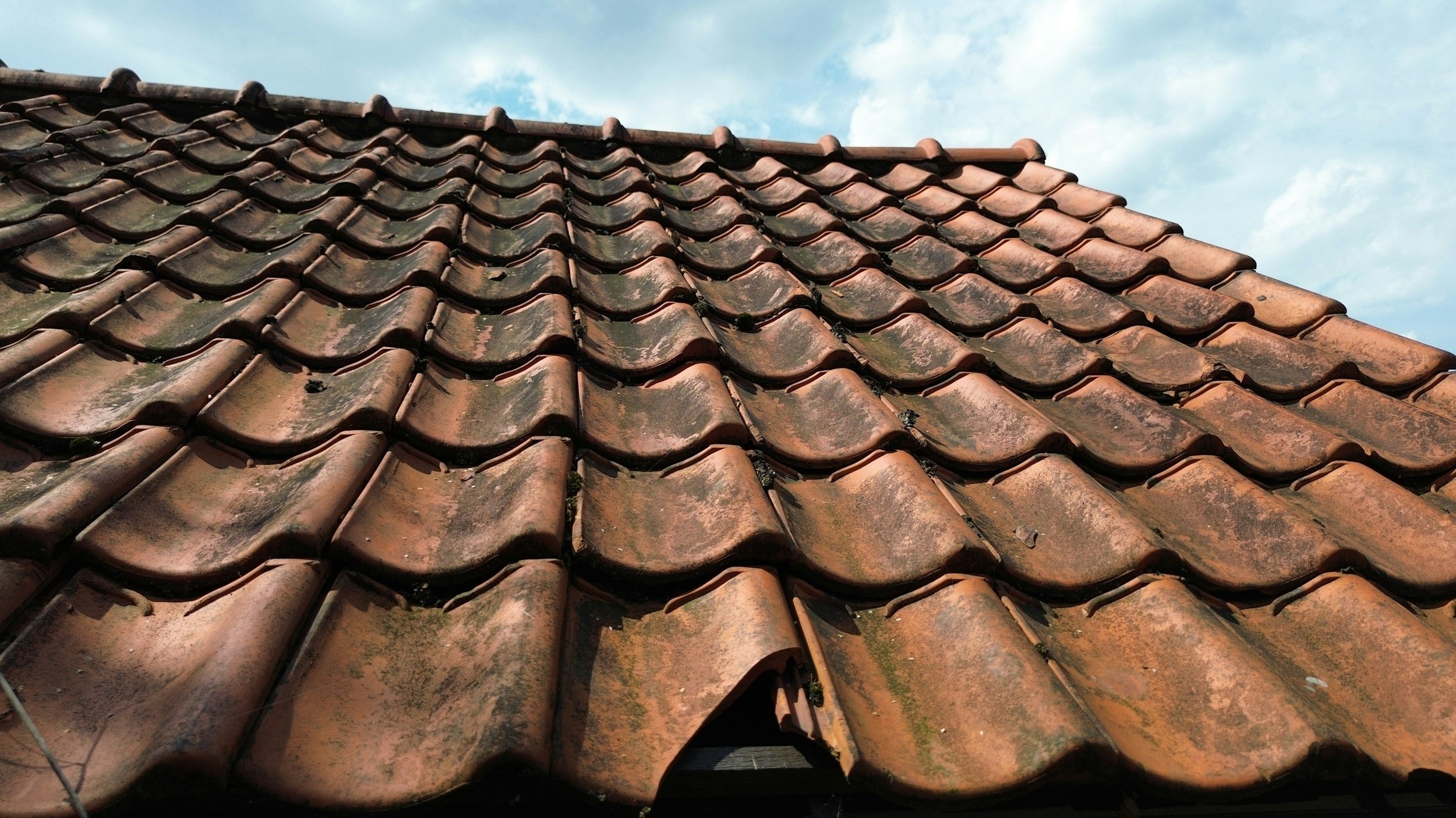Close-up of an old, weathered terracotta tiled roof with moss and dirt, under a partly cloudy sky.