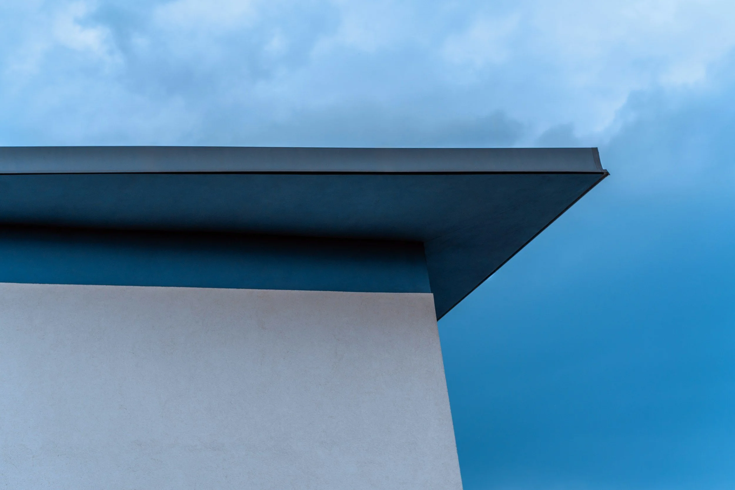 Close-up of a modern building's corner with beige, blue, and gray surfaces against a cloudy sky.