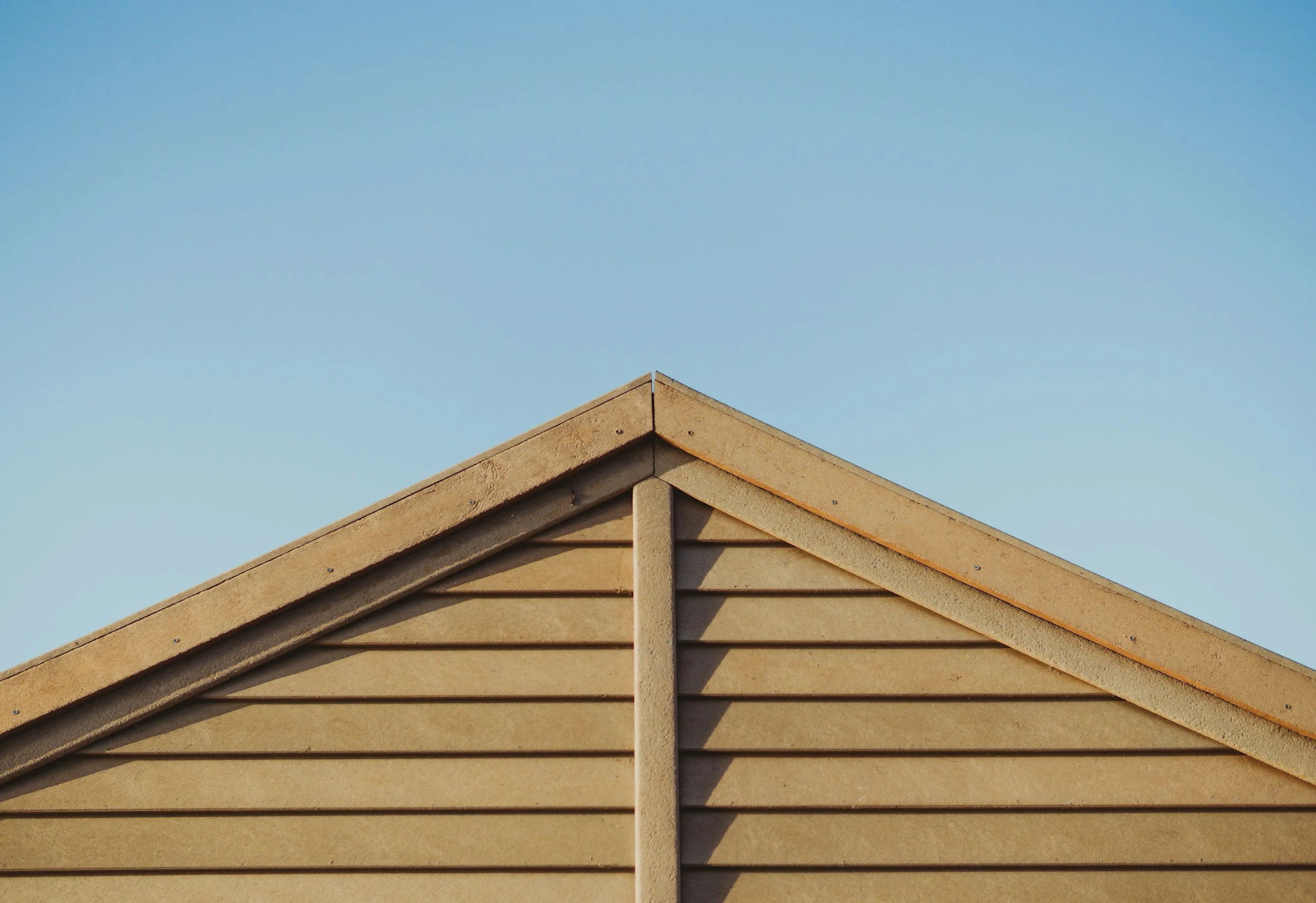 Close-up of a beige, wooden house roof with a triangular peak, against a clear blue sky.