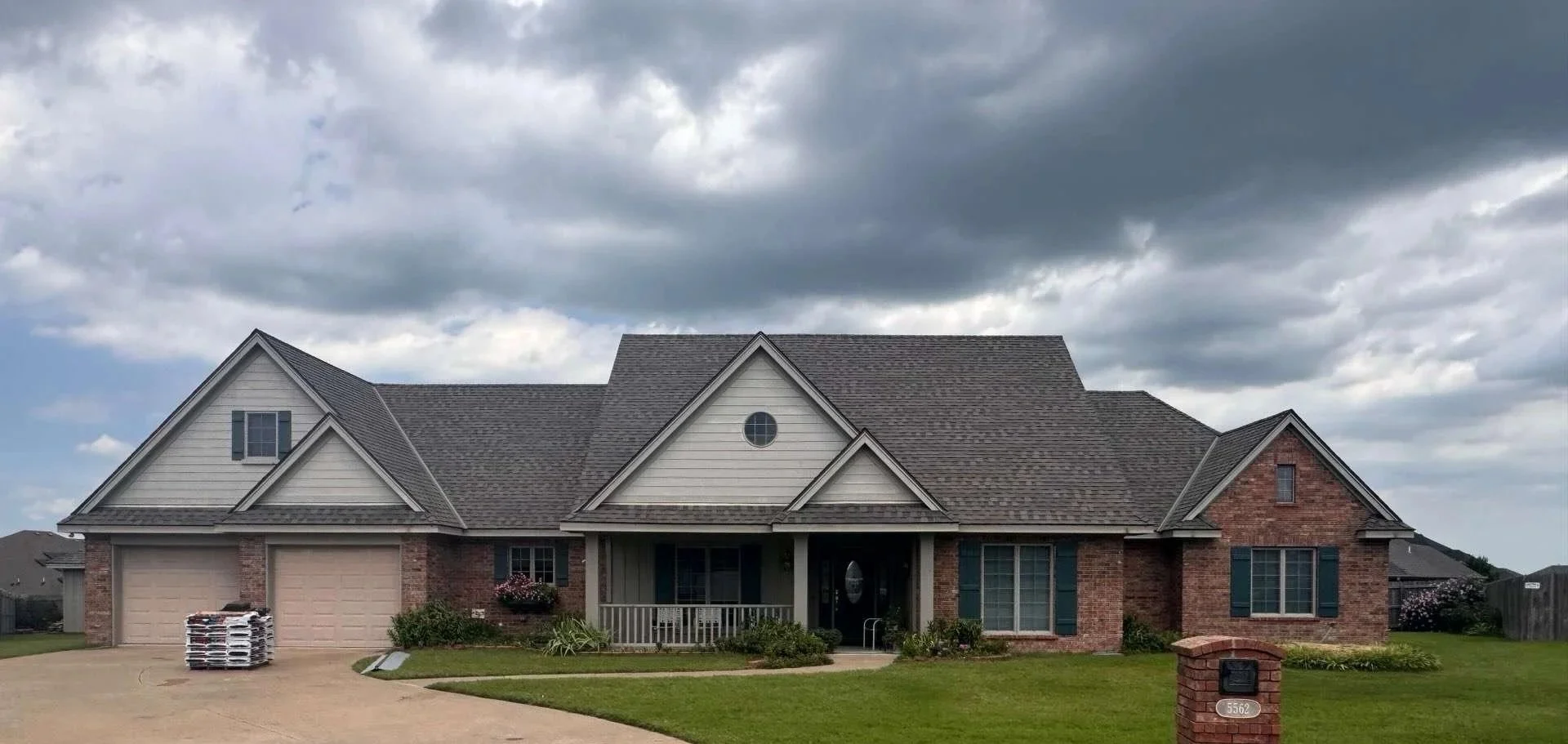 A large suburban house with a brick and siding facade, multiple gabled roof sections, a two-car garage, and a front porch with a railing, set against a dark, cloudy sky.