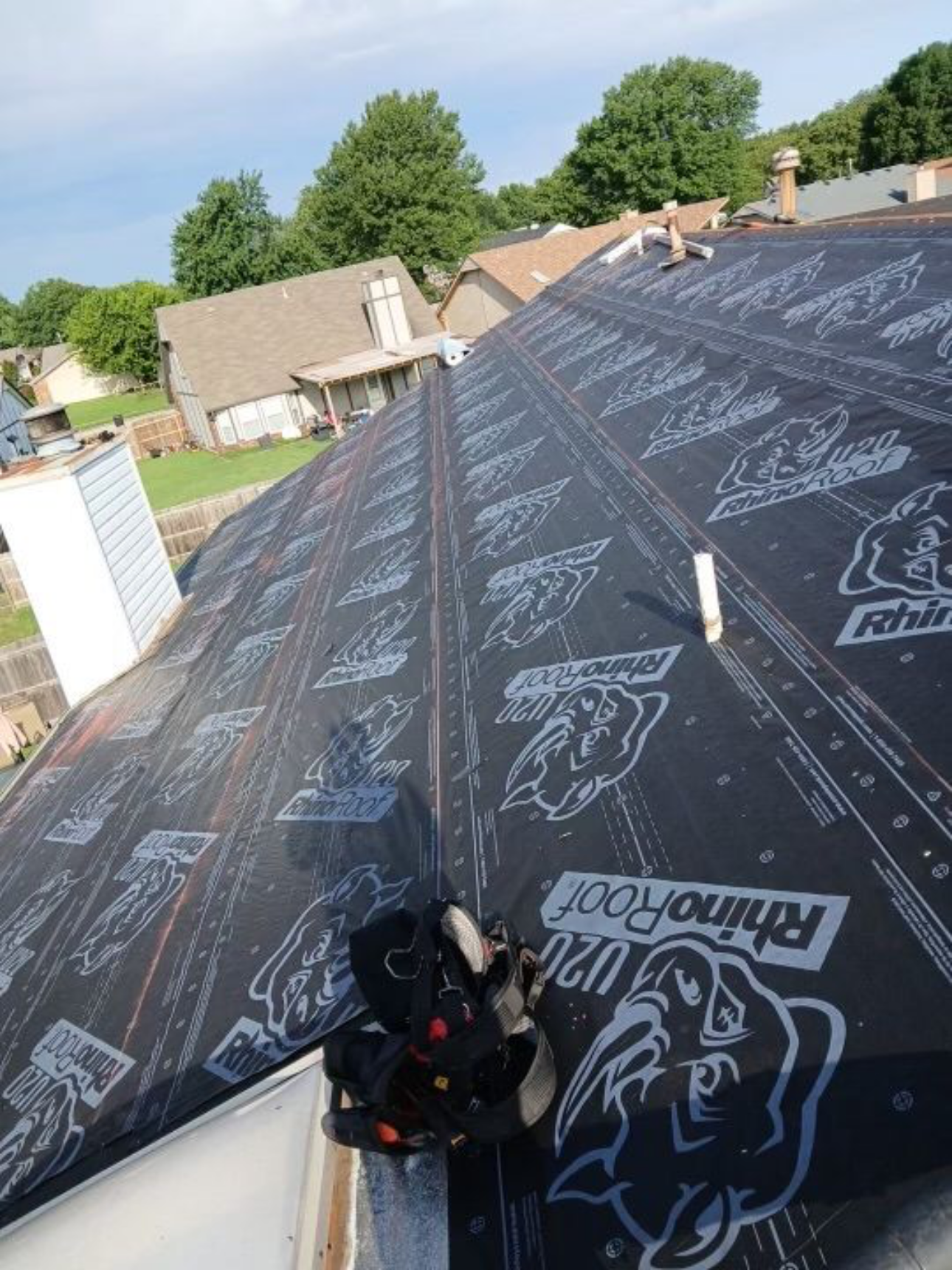 The image shows a roof under construction with a layer of weatherproofing material branded 'RhinoShield'. Several houses with trees and a blue sky are visible in the background.