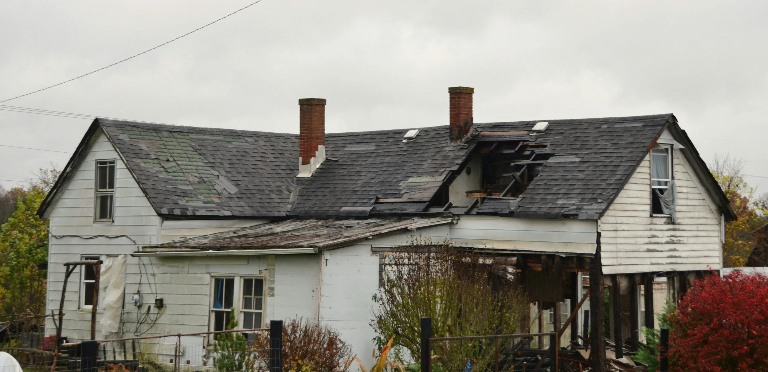 A damaged two-story house with a collapsed roof and missing shingles, showing signs of fire or structural damage, with a cloudy sky overhead.