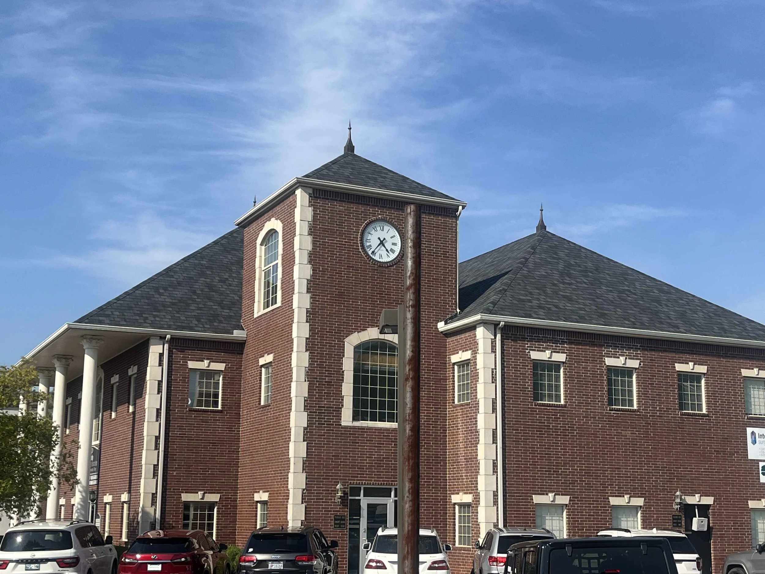 A red brick building with large white framed windows and a clock tower on top showing 10:15. The building has a dark shingled roof and white columns at the front entrance. Several cars are parked in front of the building and there is a bright blue sk