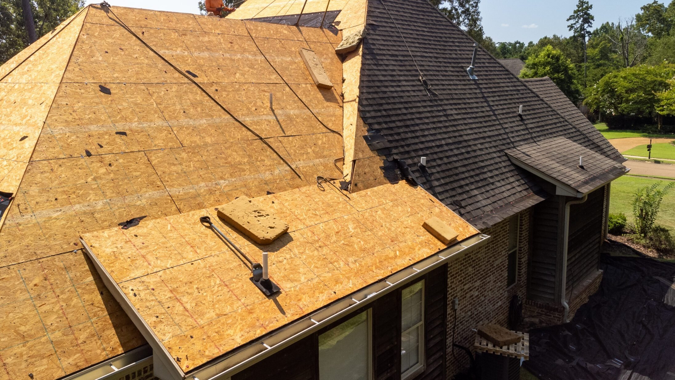 A house with a sloped roof under construction, showing exposed plywood sheathing on the left side and finished dark shingle siding on the right side, with construction tools and vents visible.