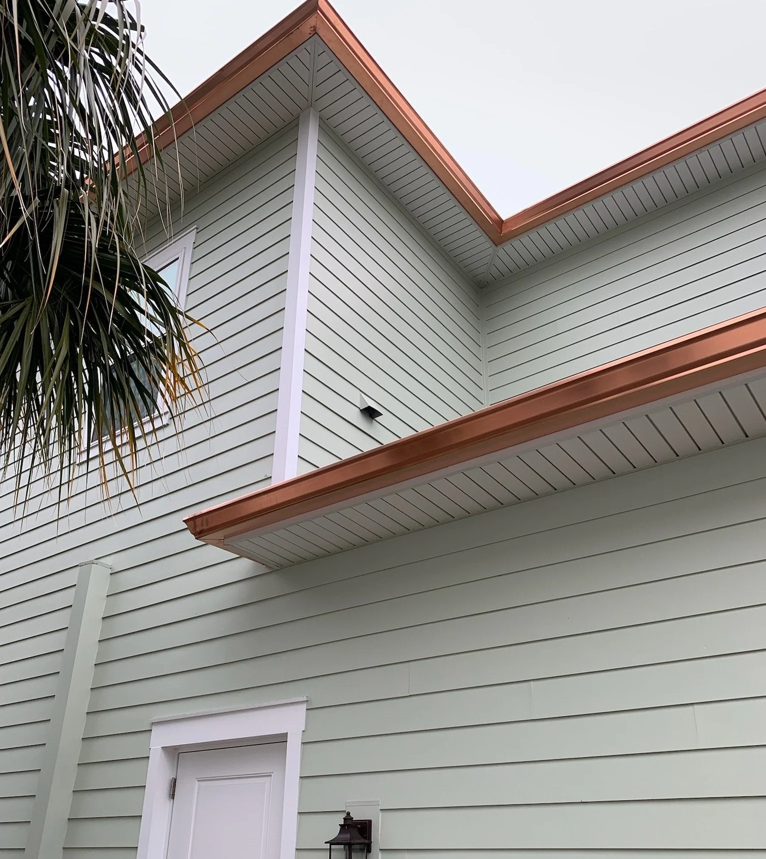 Close-up of the upper corner of a house with light green horizontal siding, a white door, a small window, a brown roof edge, and copper gutters.