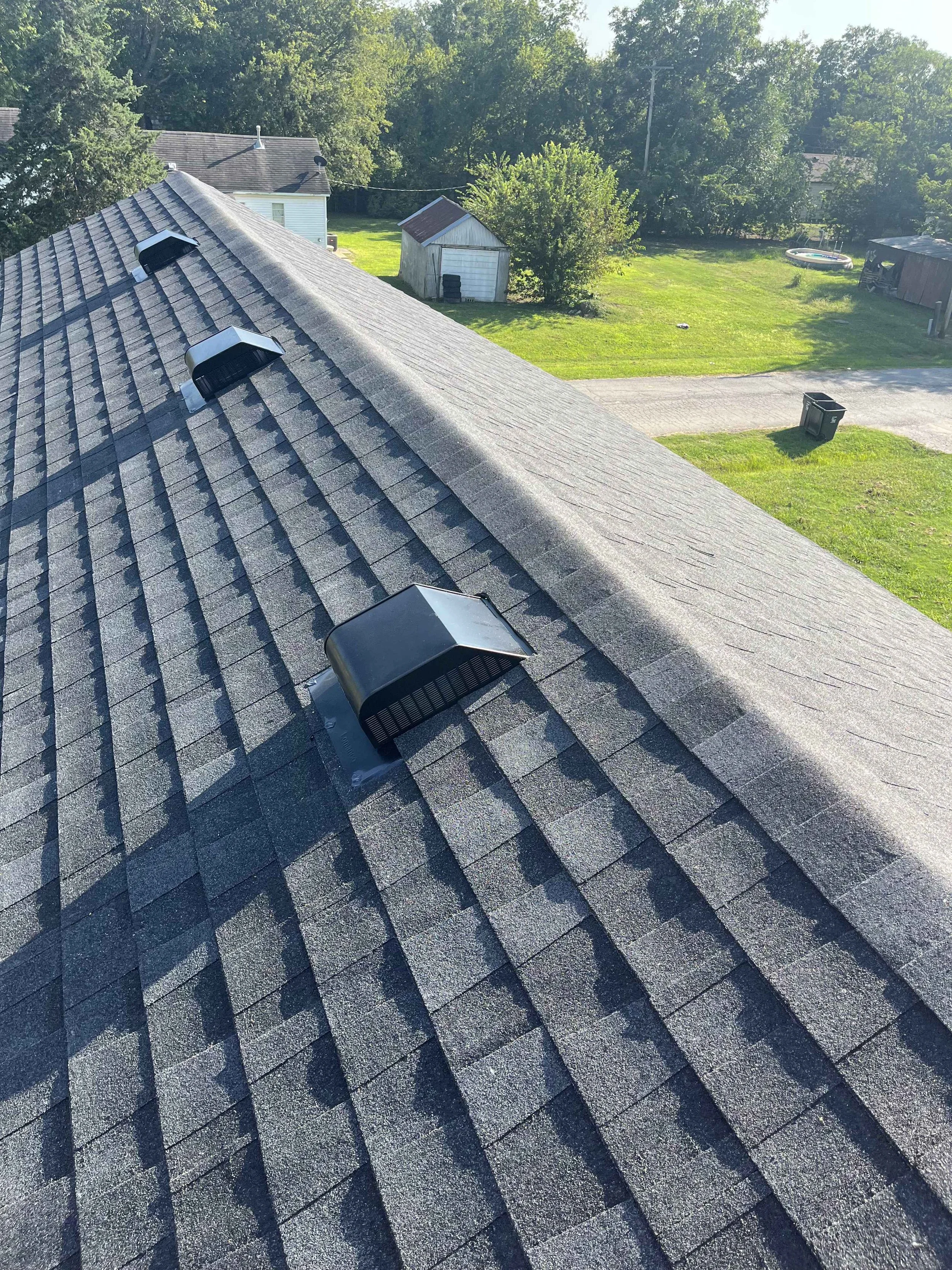 View of a rooftop with three attic vents, a green backyard with trees, a shed, a trampoline, a hot tub, and trash bins, under clear blue sky.