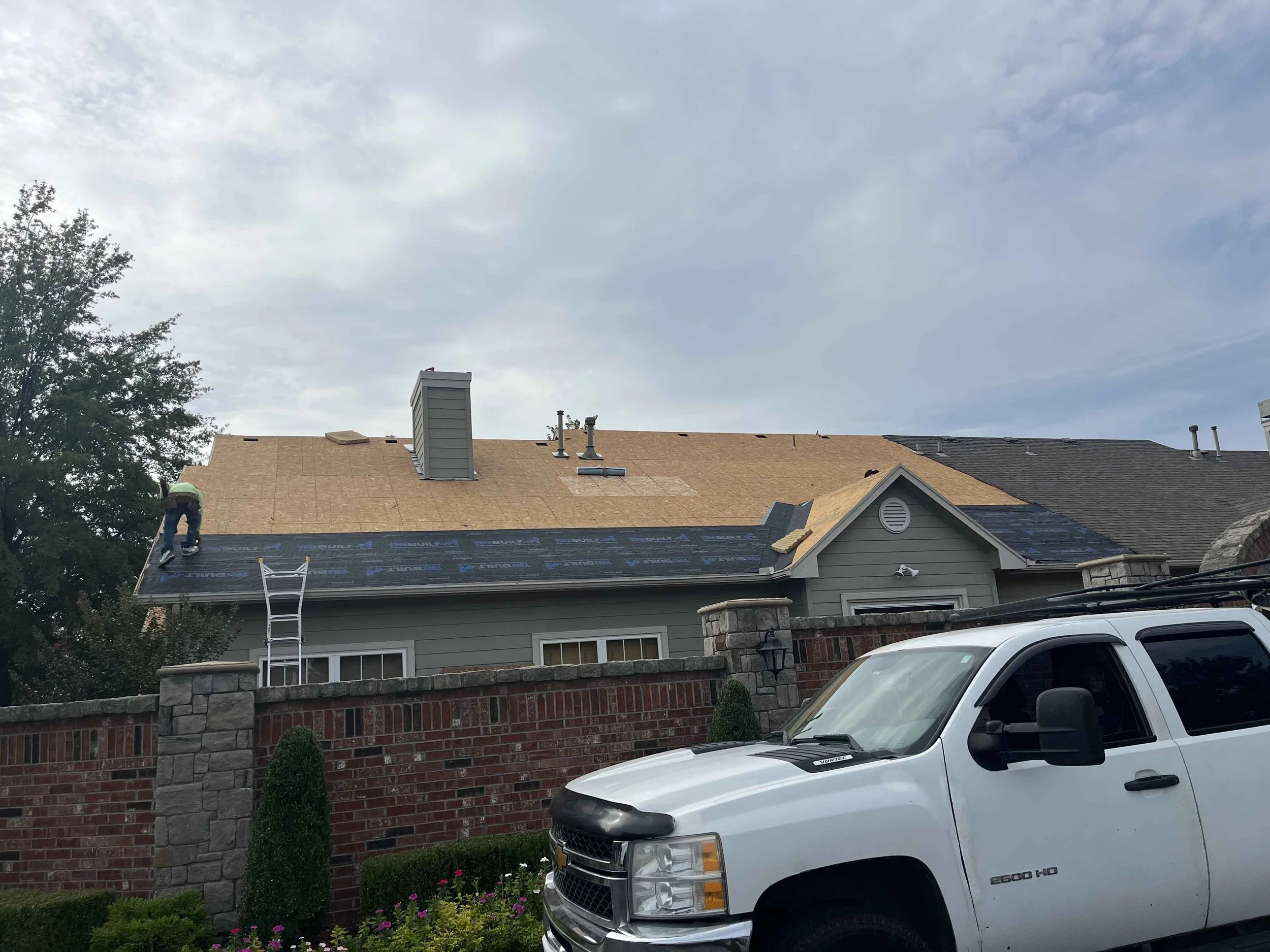 Person working on roof of house with shingles, with ladder leaning against the roof, under a cloudy sky, with a tree on the left and cars parked in front of the house.