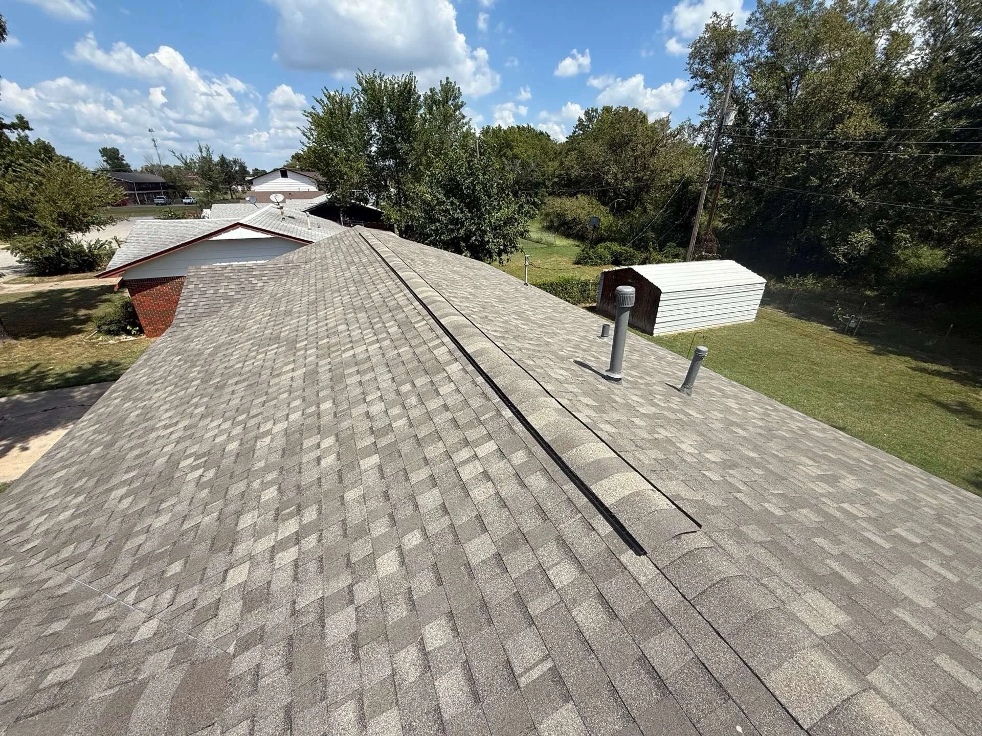 View of house rooftops with asphalt shingles, surrounded by trees, power lines, and a partly cloudy sky.