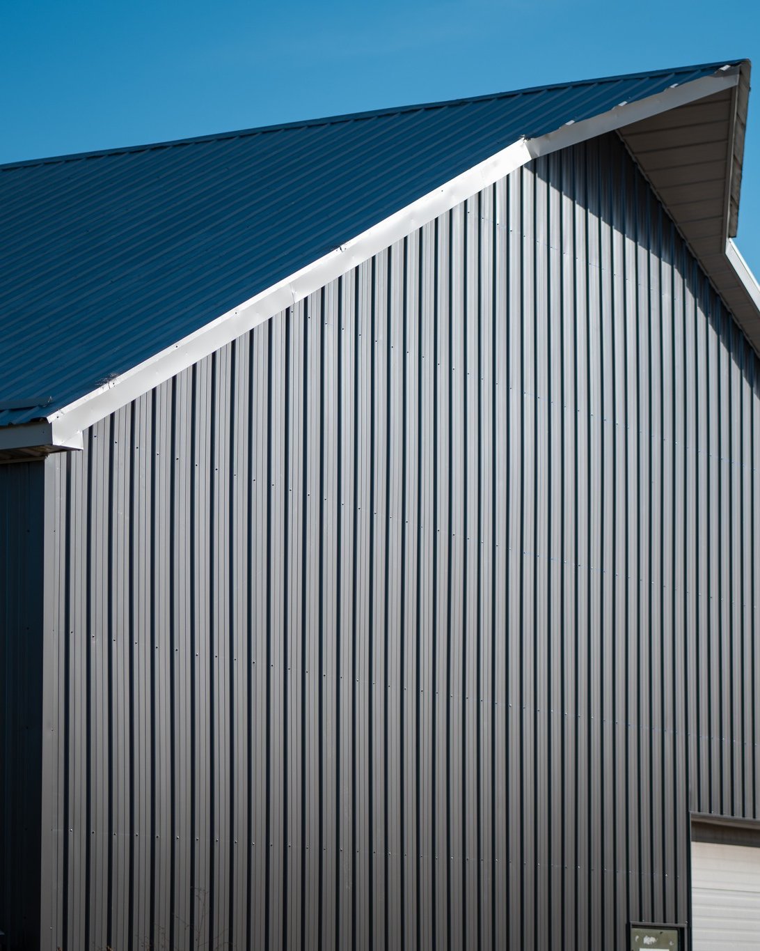 Close-up of a silver metal building with vertical corrugated panels and a blue metal roof against a clear blue sky.